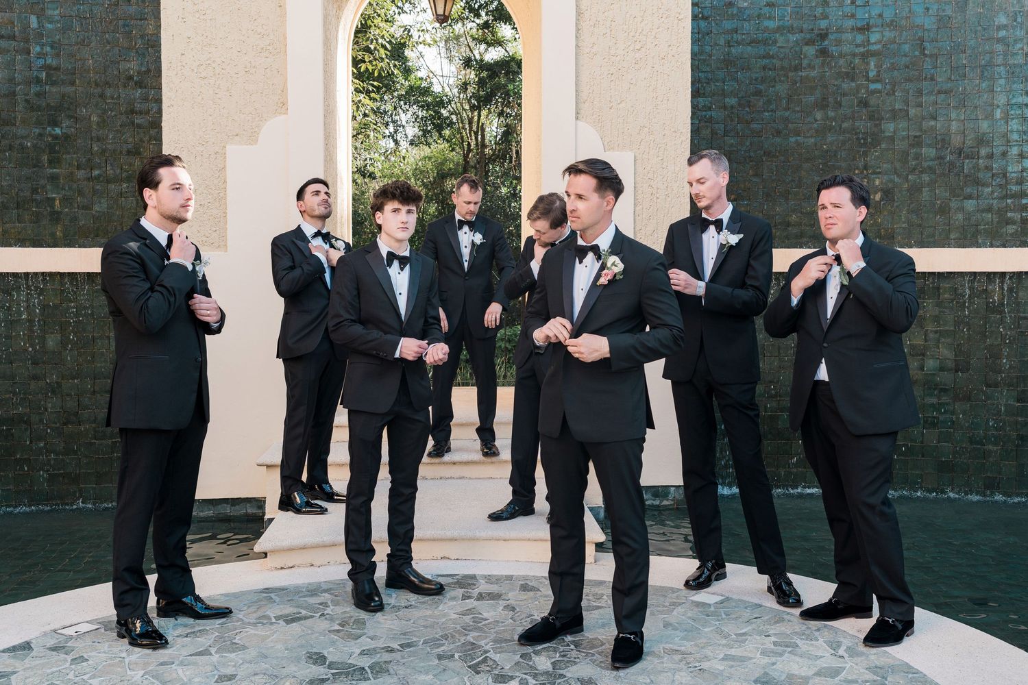 Groom and groomsmen in black tuxedos adjusting bow ties outdoors at elegant wedding venue.
