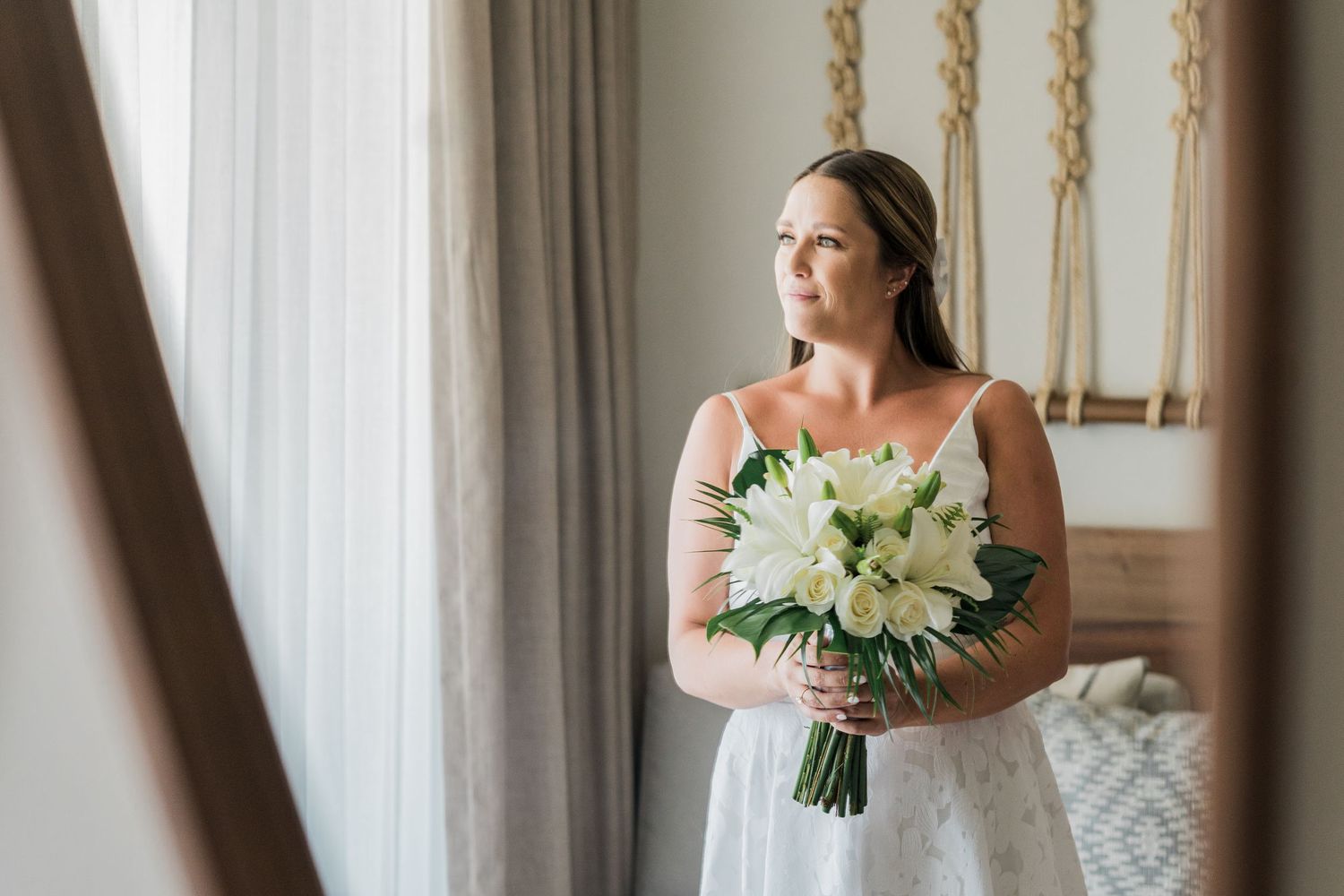 Bride in white dress holding white lily bouquet standing by window in elegant room.