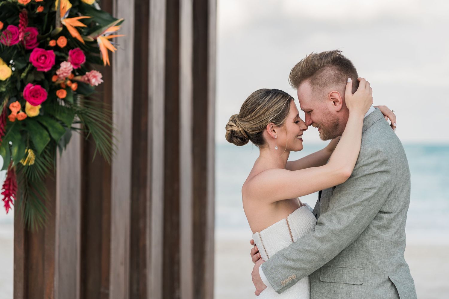 Bride and groom share an intimate moment embracing on a beach wedding with tropical floral arrangement backdrop.