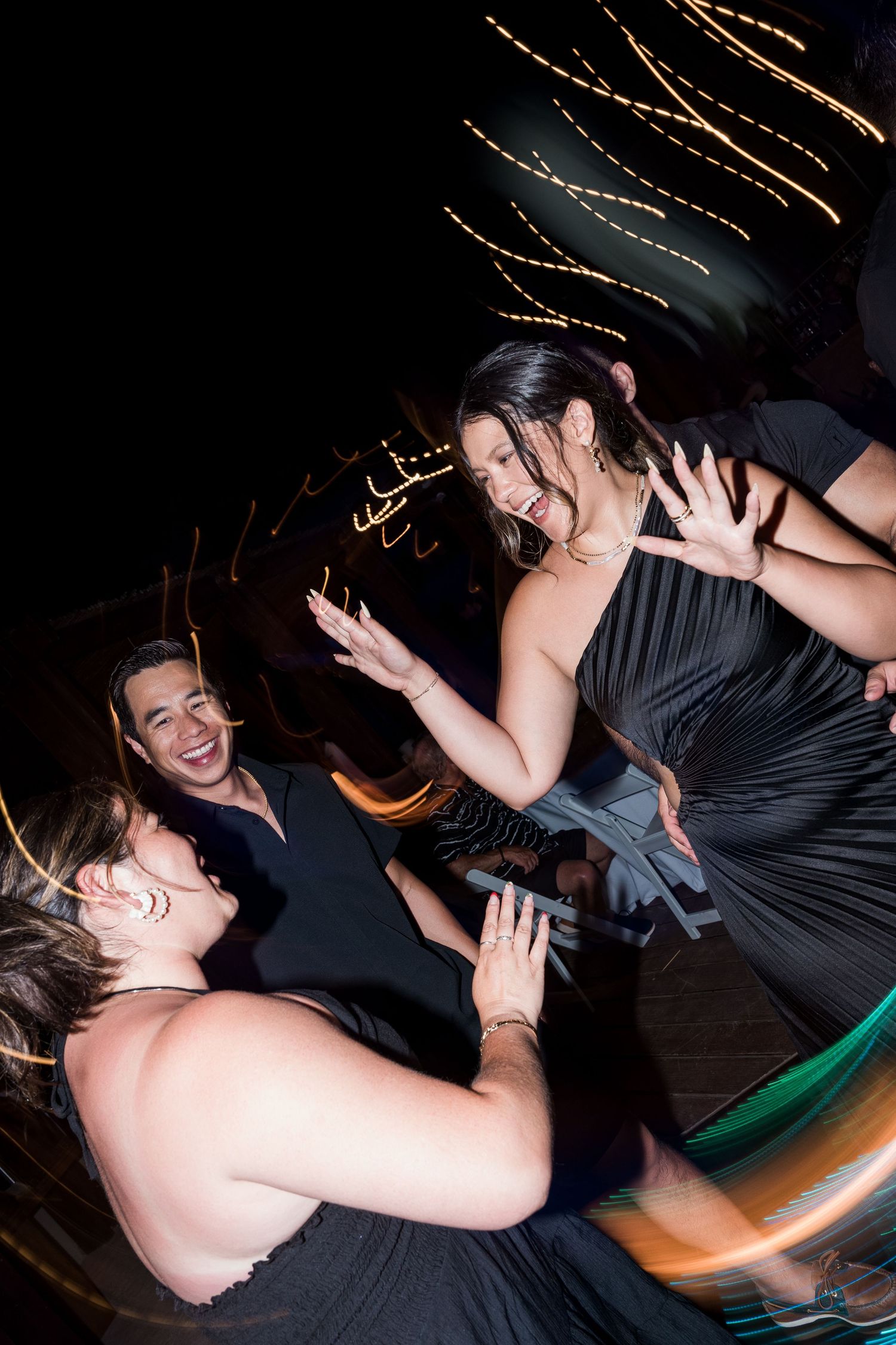 Three guests in formal attire laugh and dance together at a nighttime event with festive lights.