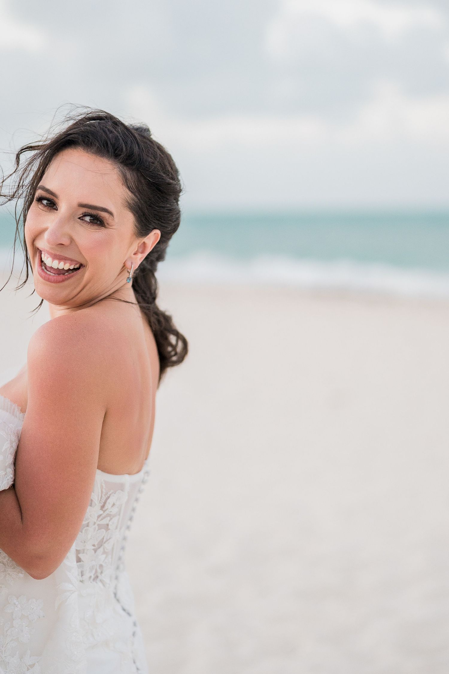 Smiling bride in a white lace strapless gown poses on a sandy beach with ocean waves in the background.
