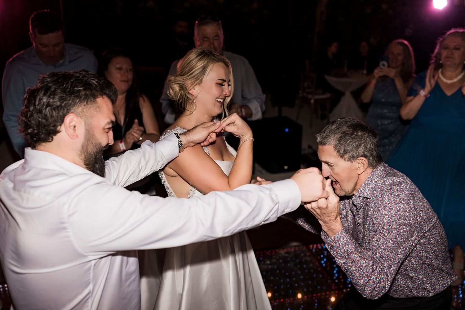 Groom kisses guest's hand while bride laughs joyfully during lively wedding reception dance floor celebration.