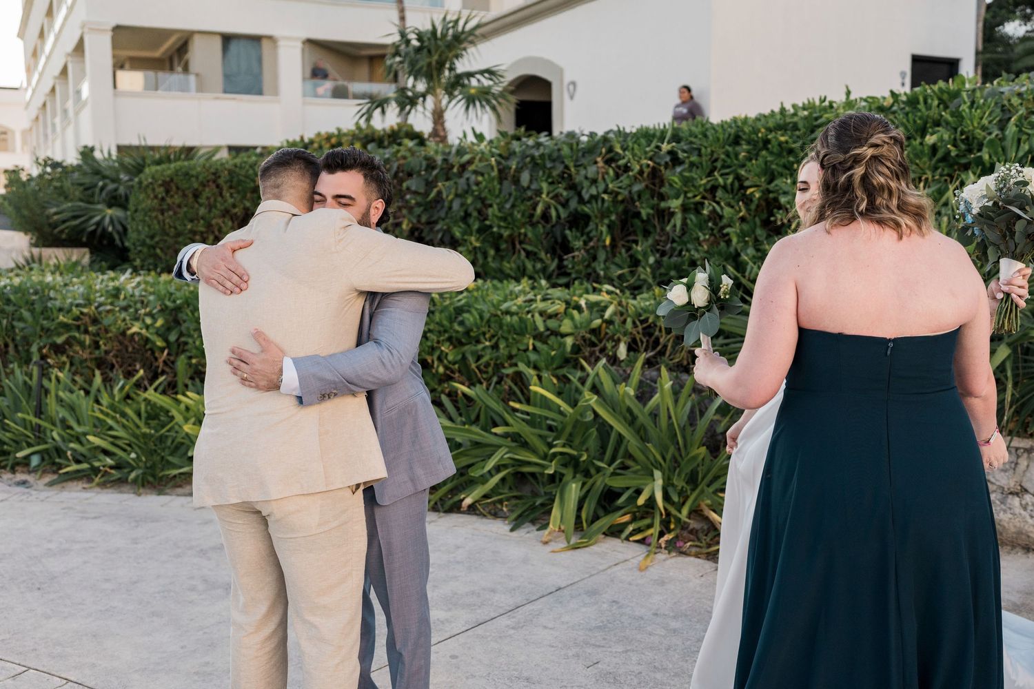 Two men in suits share a warm hug outdoors at a wedding, while a woman in a green gown stands nearby.