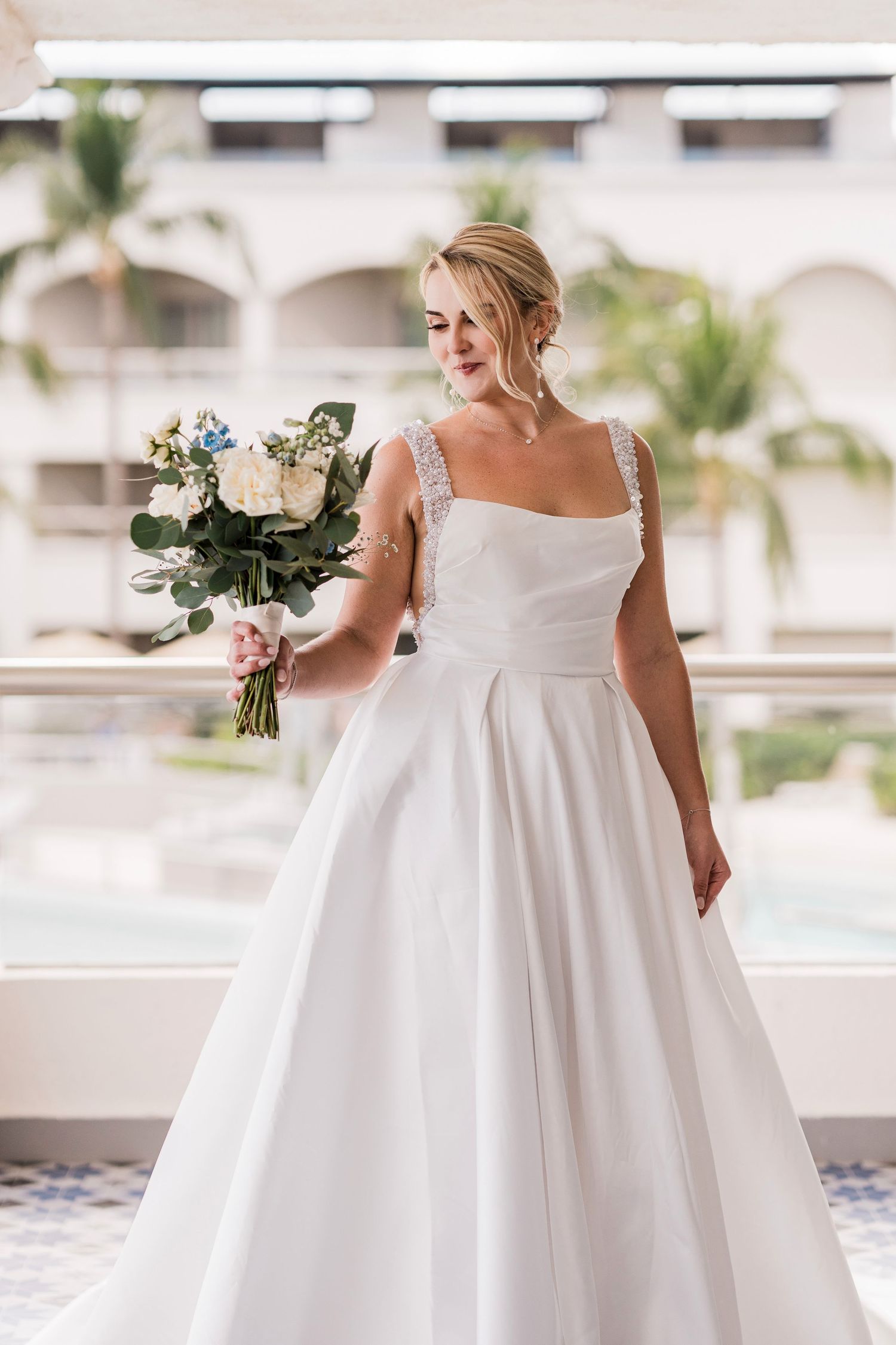 Bride in white ball gown with embellished straps holds floral bouquet outdoors near palm trees.