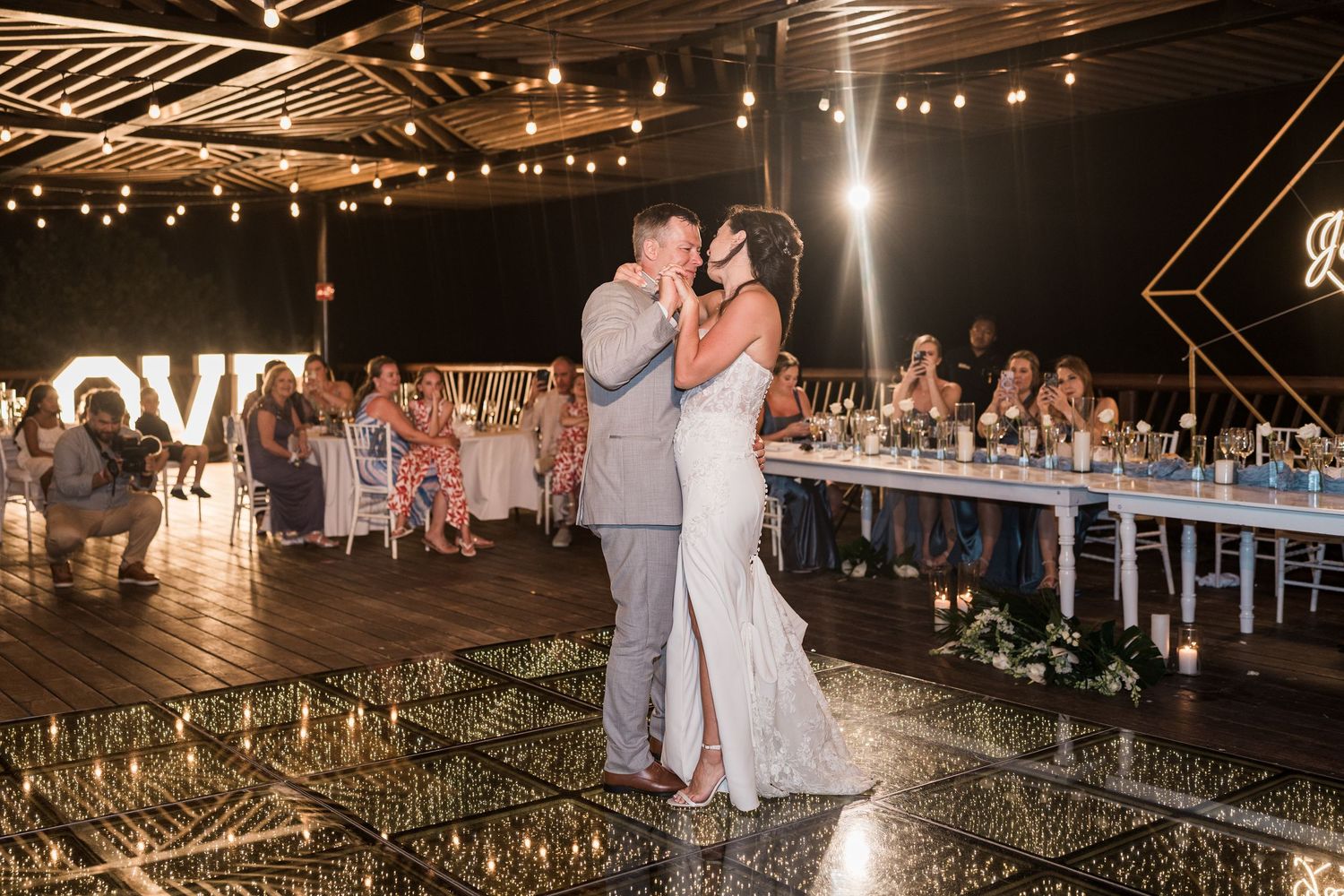 Bride and groom share first dance on mirrored floor at elegant outdoor wedding reception with string lights and LOVE sign.
