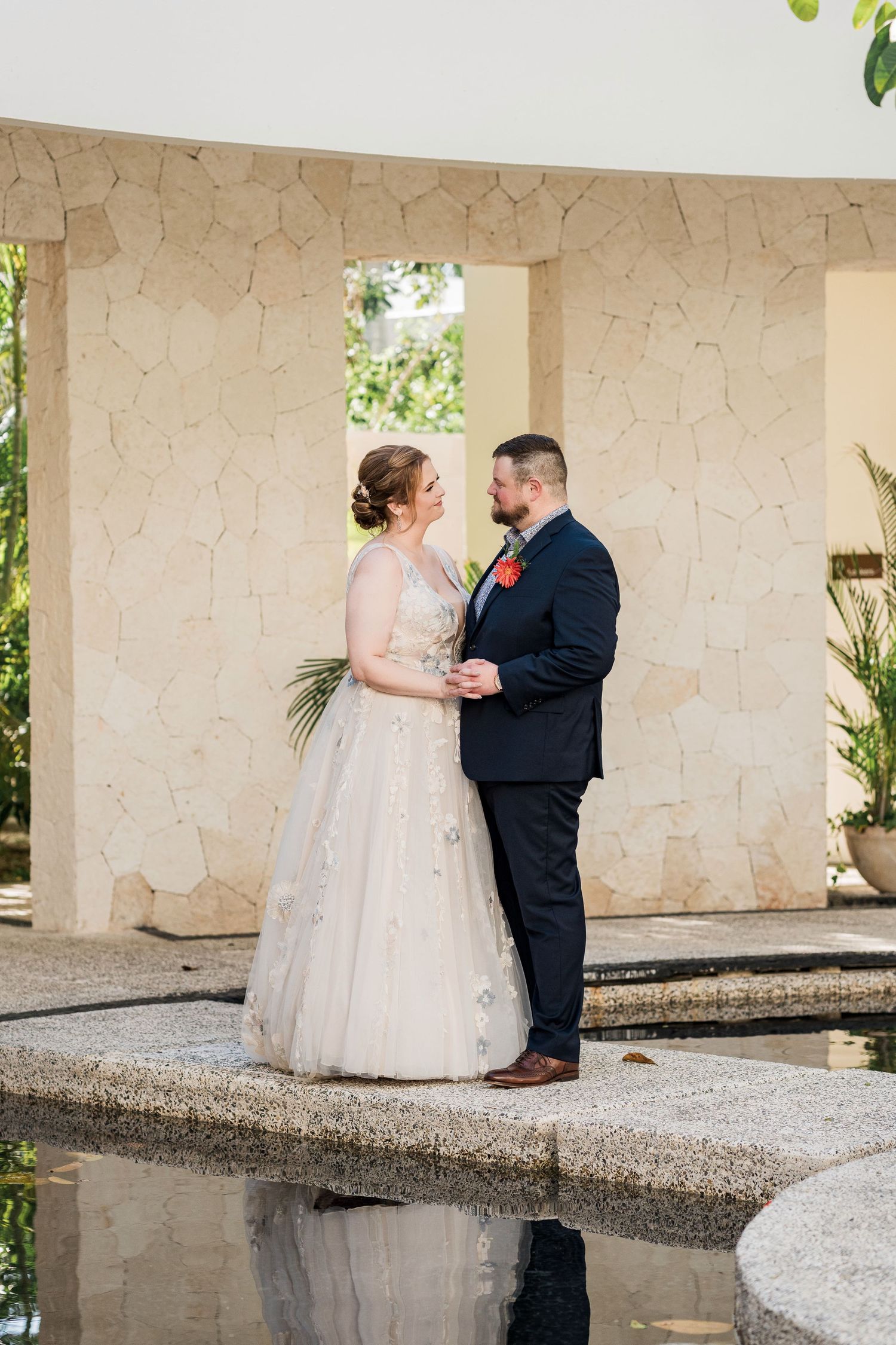 Secrets Akumal Wedding - Bride in blush gown and groom in navy suit stand facing each other near a reflecting pool