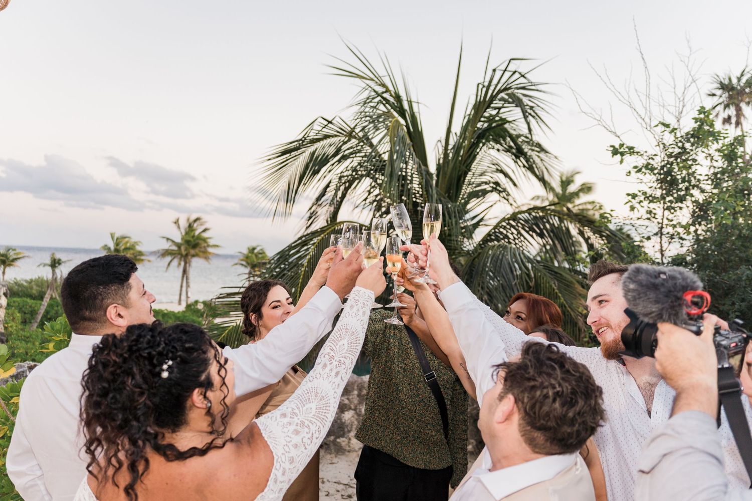 Xcaret Arte wedding party toasting with champagne glasses outdoors near tropical palm trees at a beach celebration.