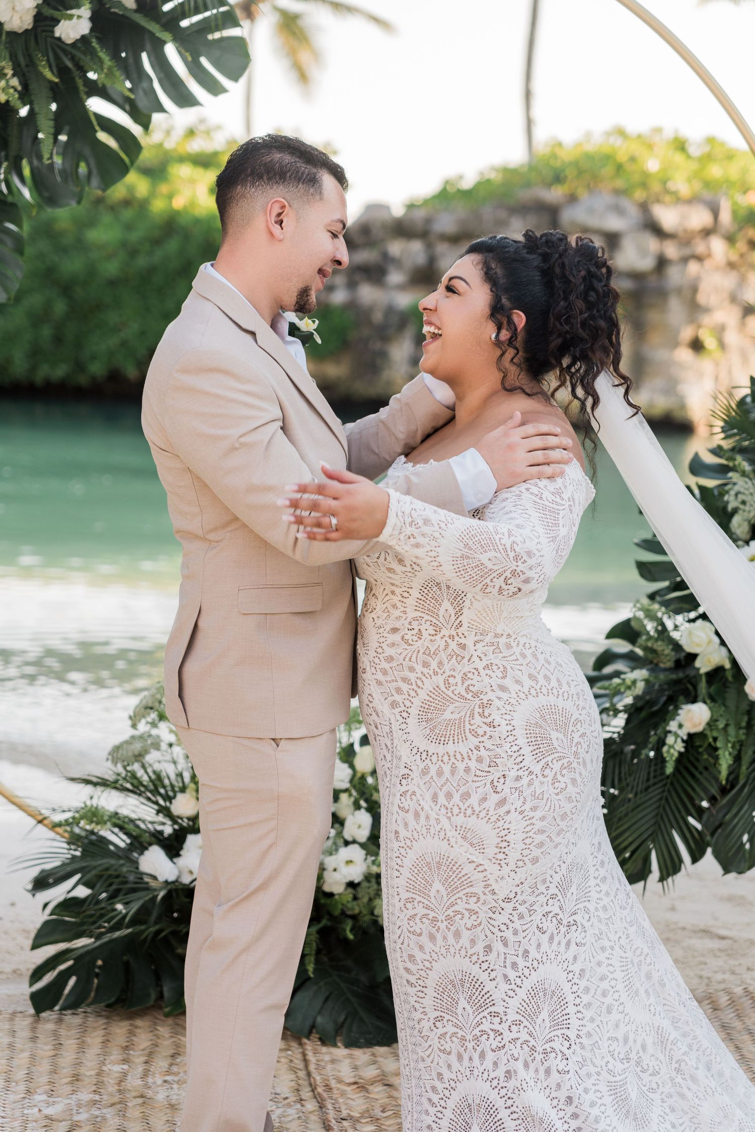 Xcaret Arte Wedding - Bride and groom share a joyful moment at a tropical beach wedding ceremony with floral arch backdrop.