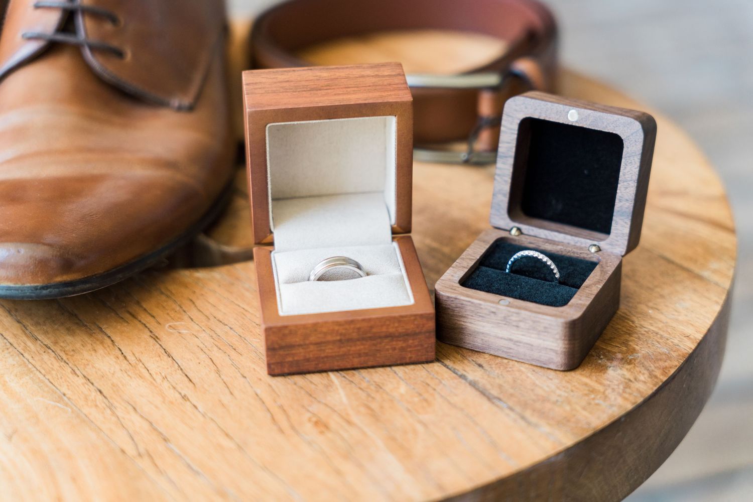 Xcaret Arte Wedding - Two wooden wedding ring boxes with gold bands displayed on a wooden surface beside brown leather dress shoes.