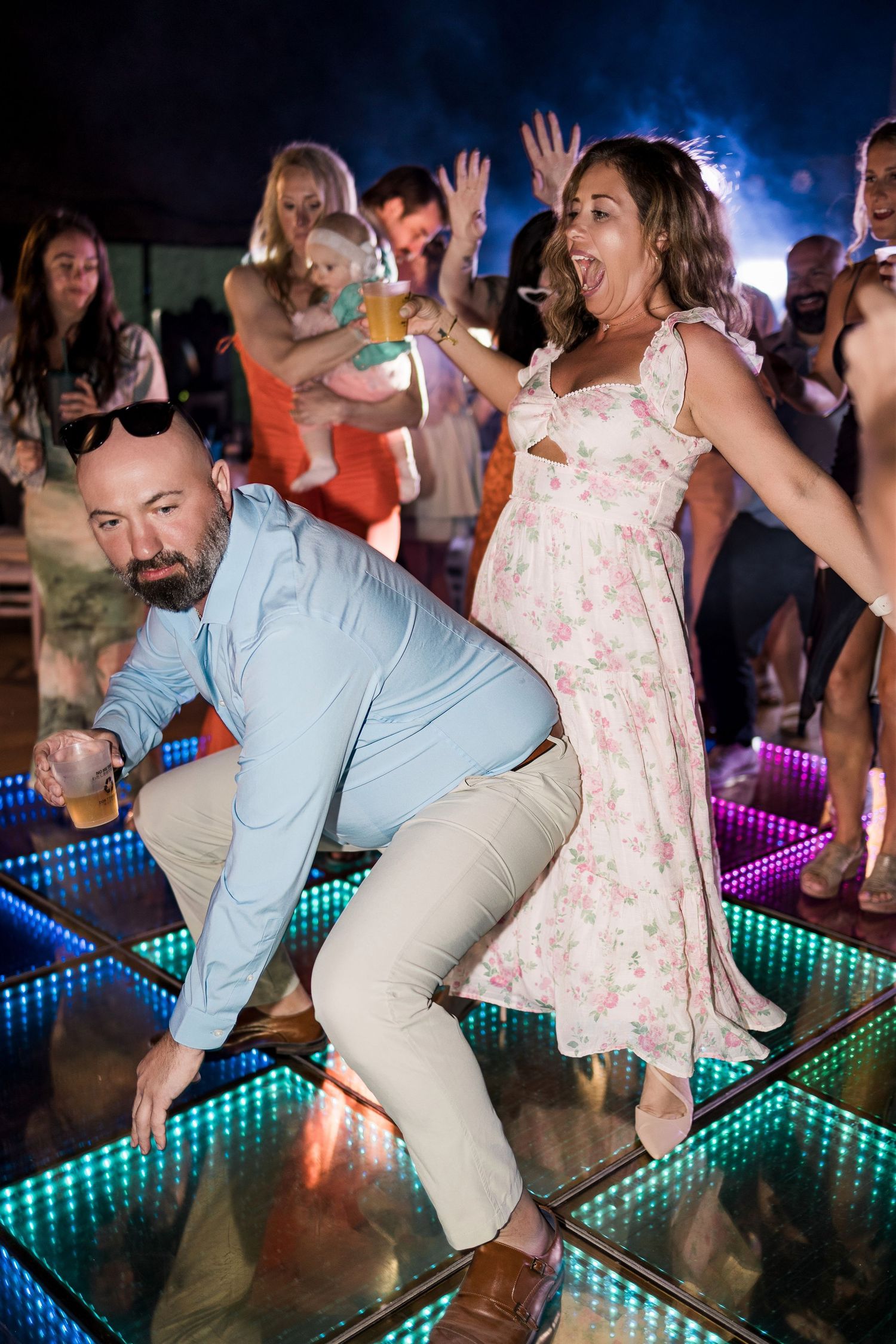 Couple dancing energetically on illuminated LED dance floor at outdoor evening party with guests celebrating behind them.