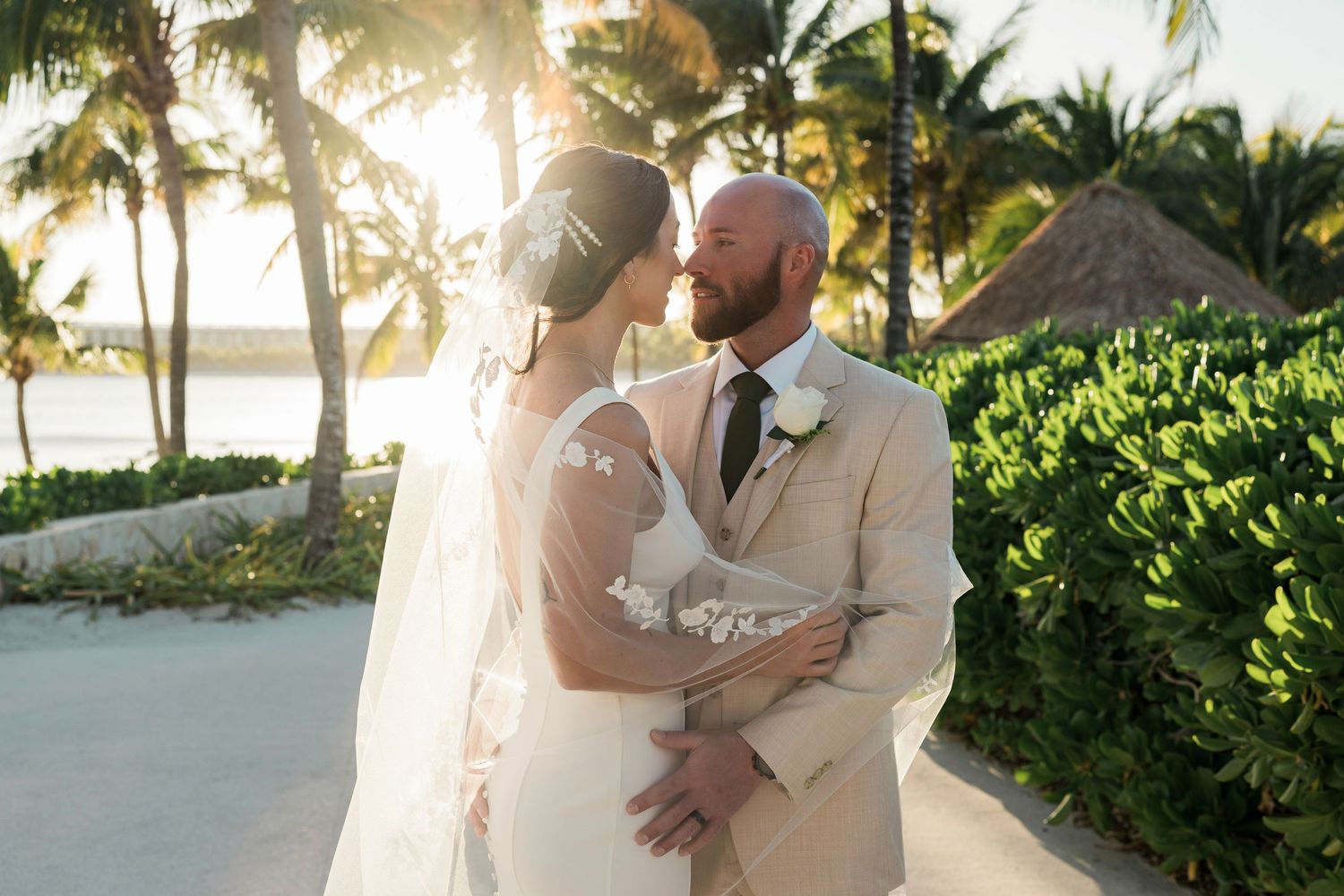 Bride and groom share a romantic moment at sunset on a tropical beach, surrounded by palm trees.