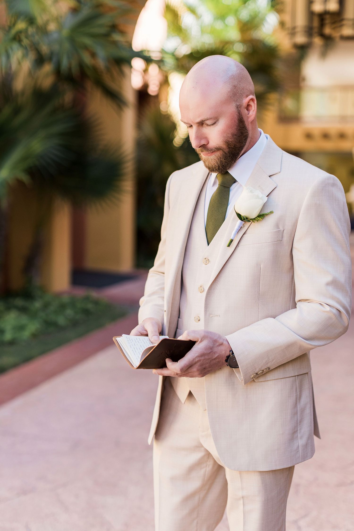 Bald groom with beard in cream suit and olive tie reads from a book outdoors, with a white rose boutonniere.
