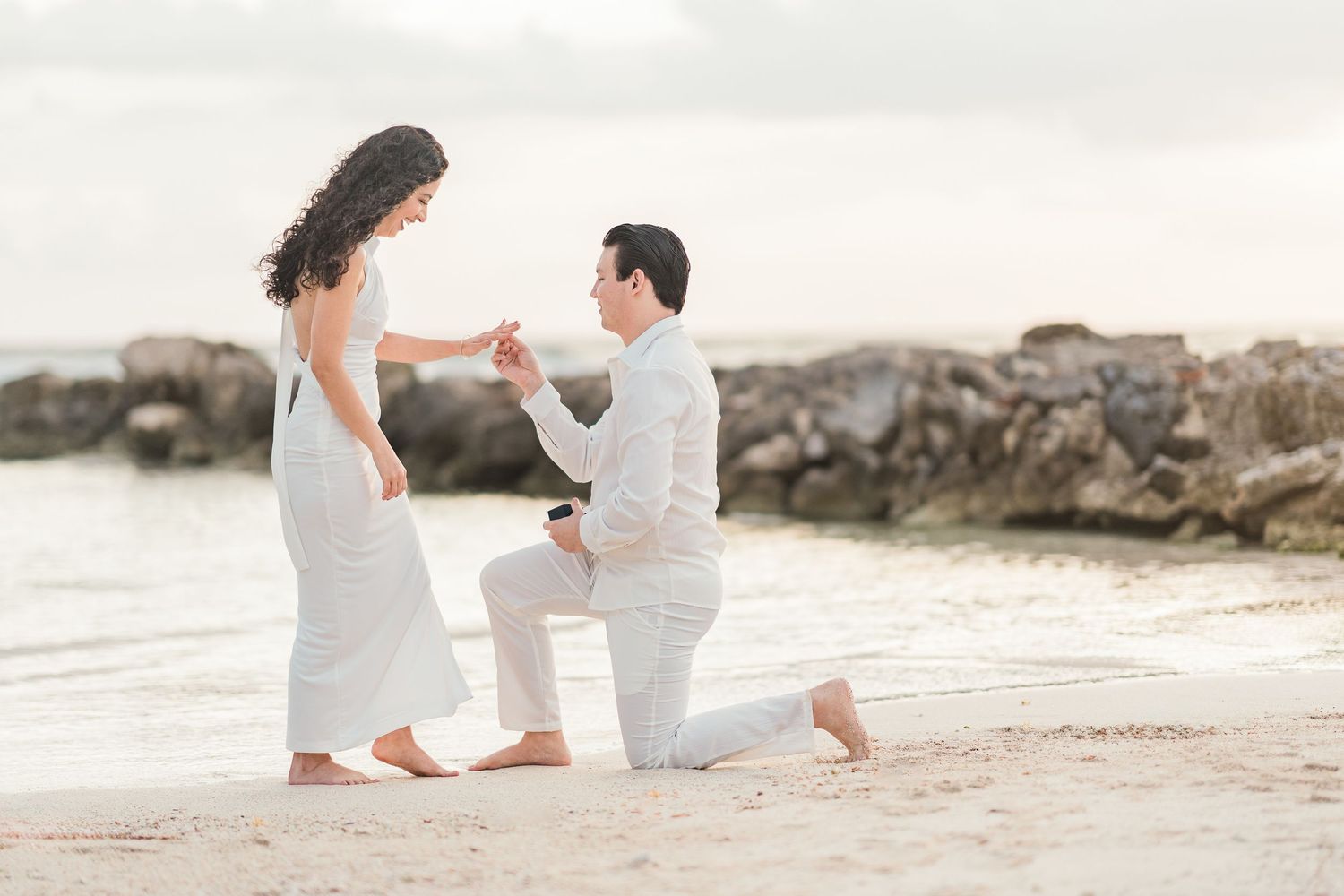Man proposing to woman on a sandy beach at sunset, both dressed in white, with rocky shoreline in the background.