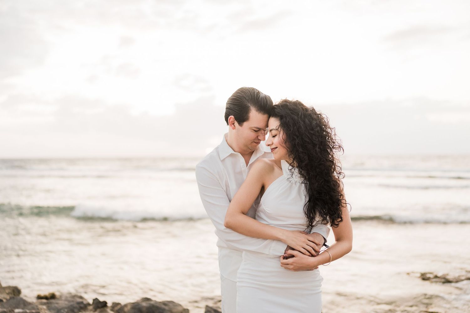 Couple embracing tenderly on a rocky beach, woman in white dress, man in white shirt, ocean waves in background.