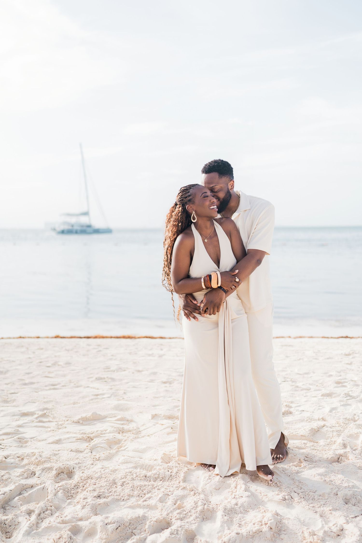 A happy Black couple in white outfits cuddles on a beach with a sailboat behind them.