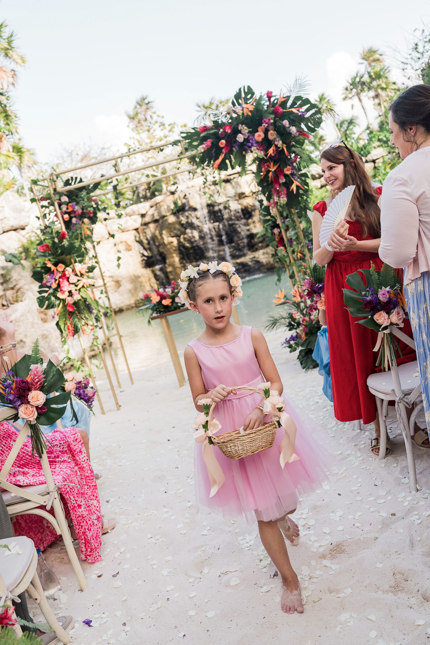 Young flower girl in pink dress walks down floral wedding aisle carrying wicker basket at tropical outdoor ceremony.