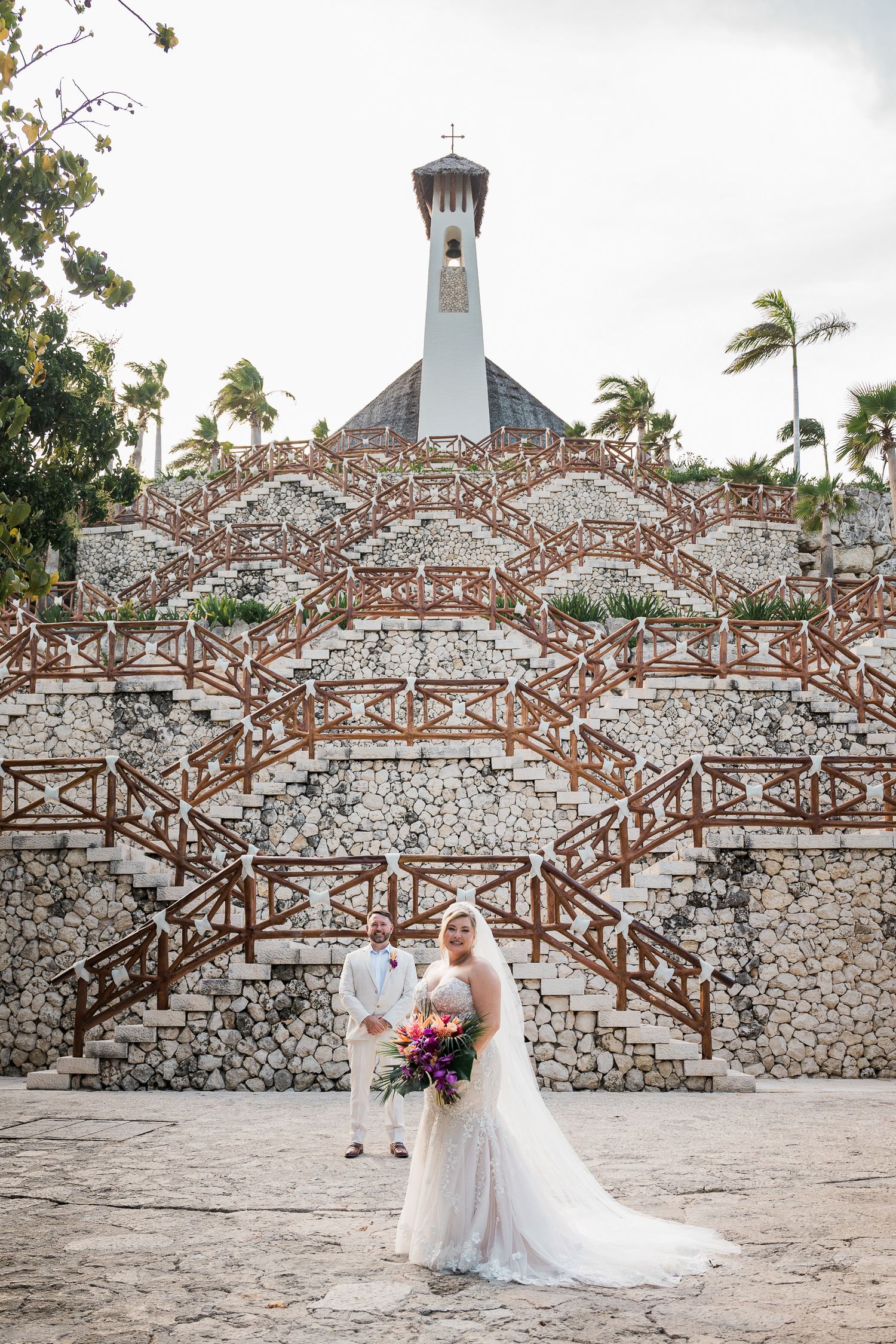 Bride and groom pose before a grand stone staircase with a chapel tower at a tropical wedding venue.
