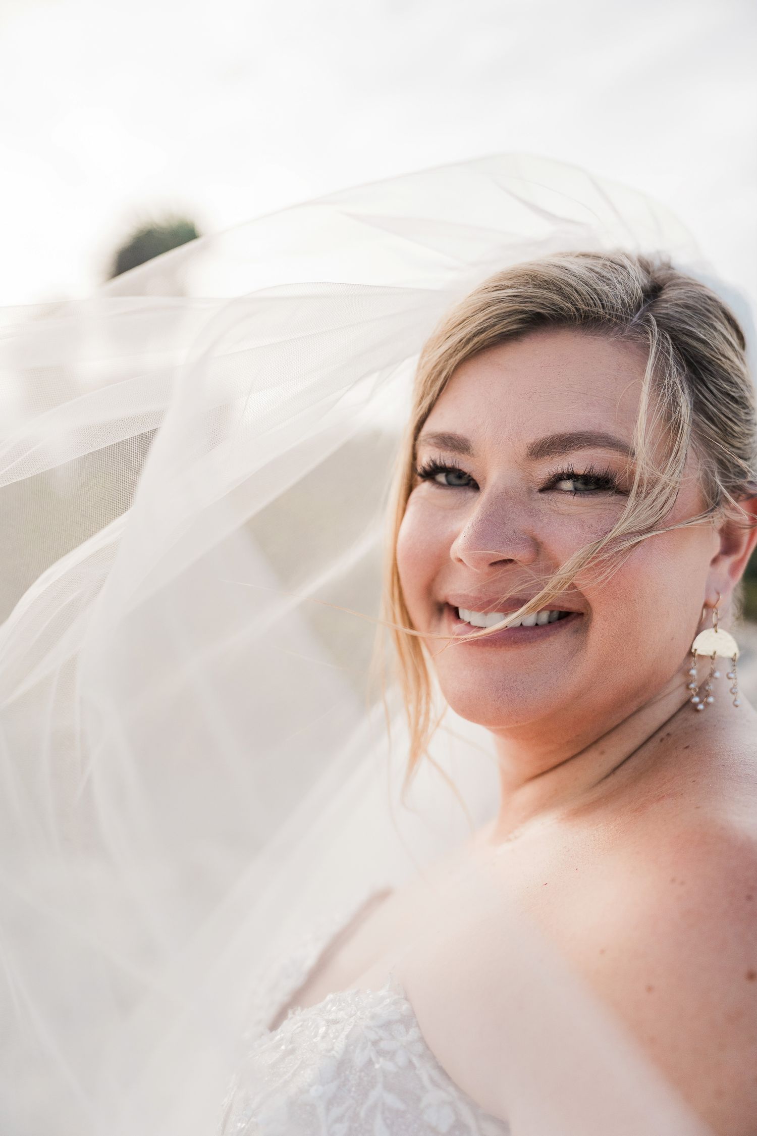 Hotel Xcaret México Wedding. Smiling bride with flowing white veil caught in wind, wearing lace dress and pearl earrings on her wedding day.