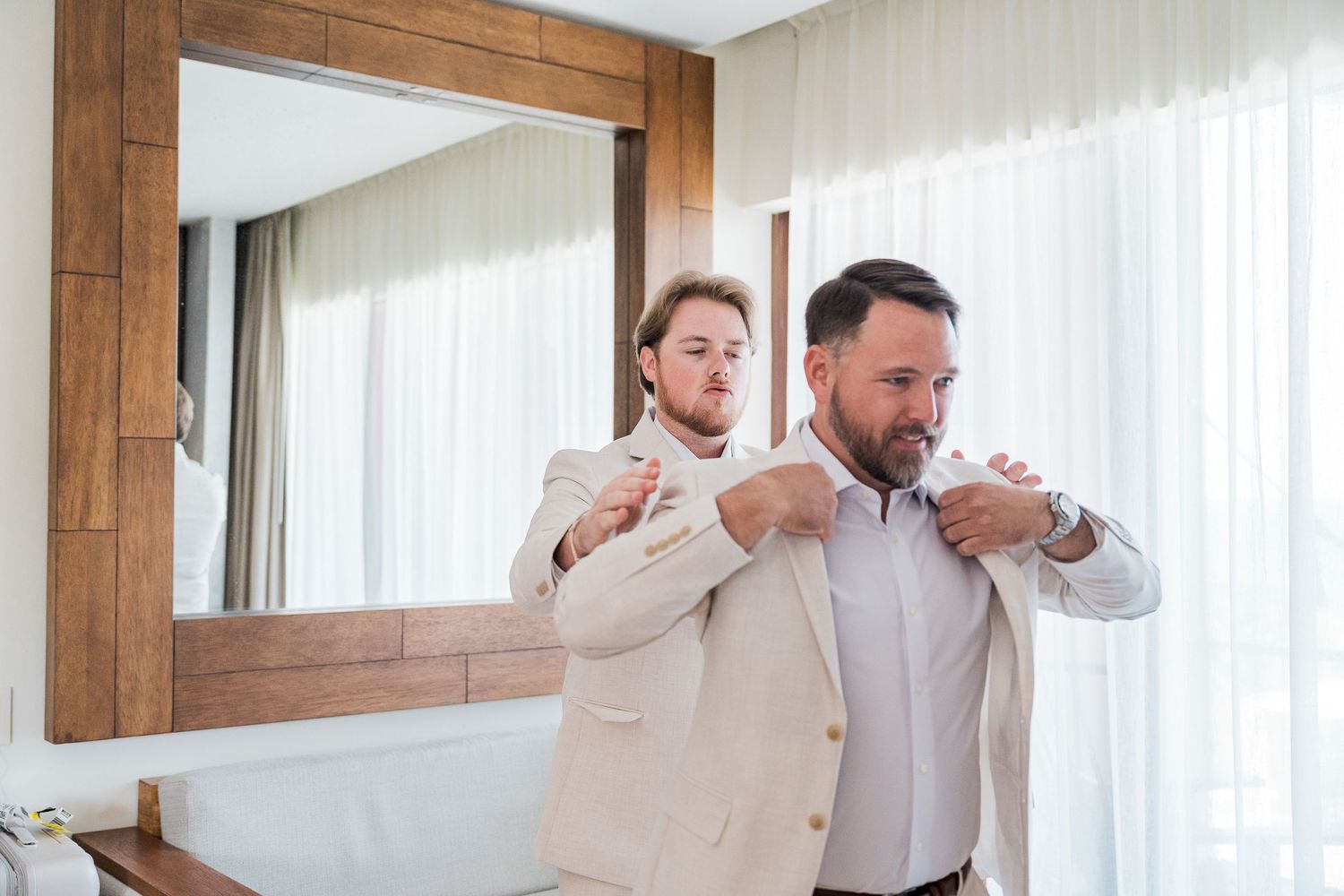 Two men in beige suits preparing for a wedding, one helping adjust the other's jacket collar in an elegant hotel room.