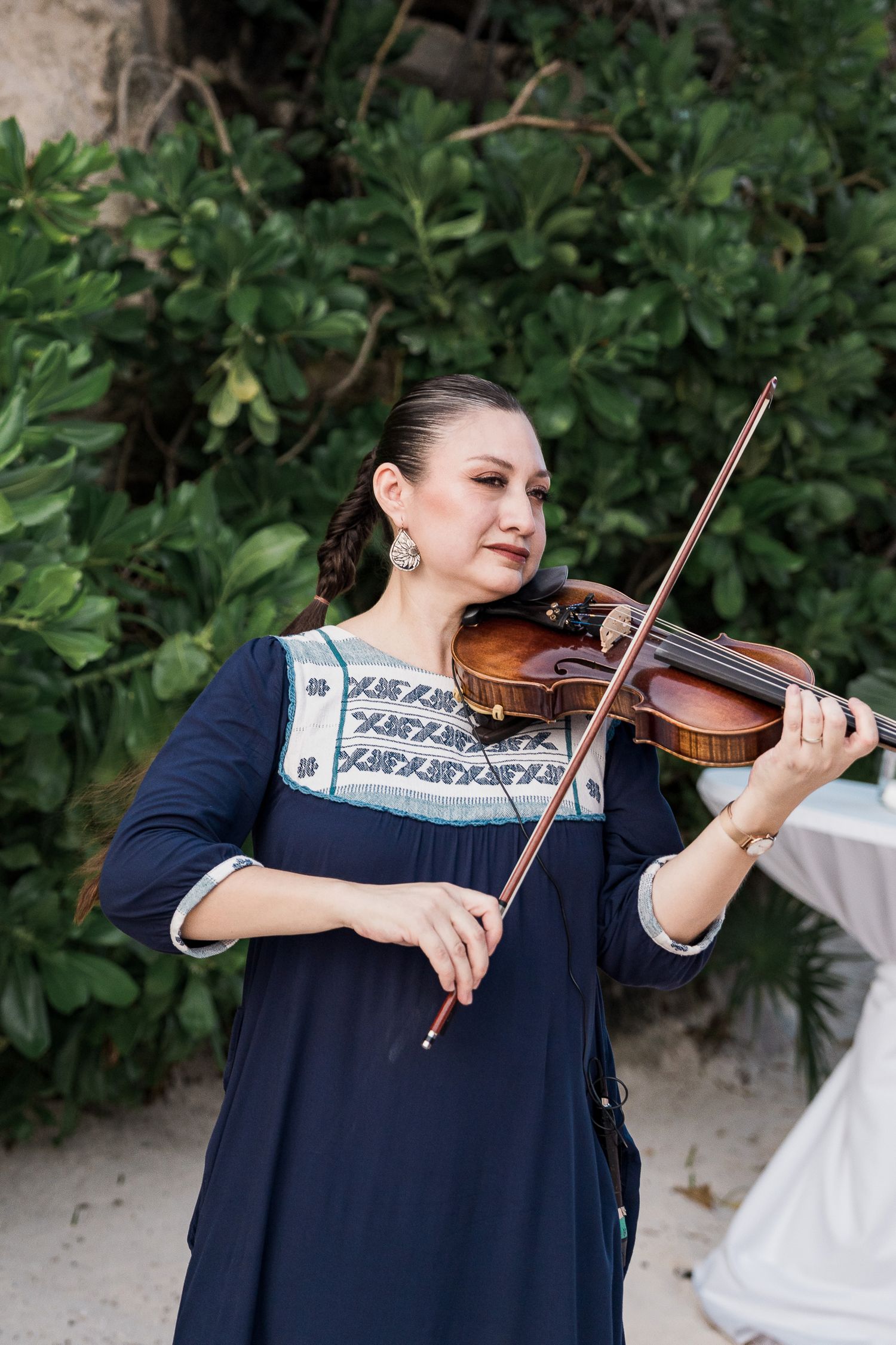 Hotel Xcaret México Woman in navy embroidered dress playing violin outdoors with lush green foliage in the background.