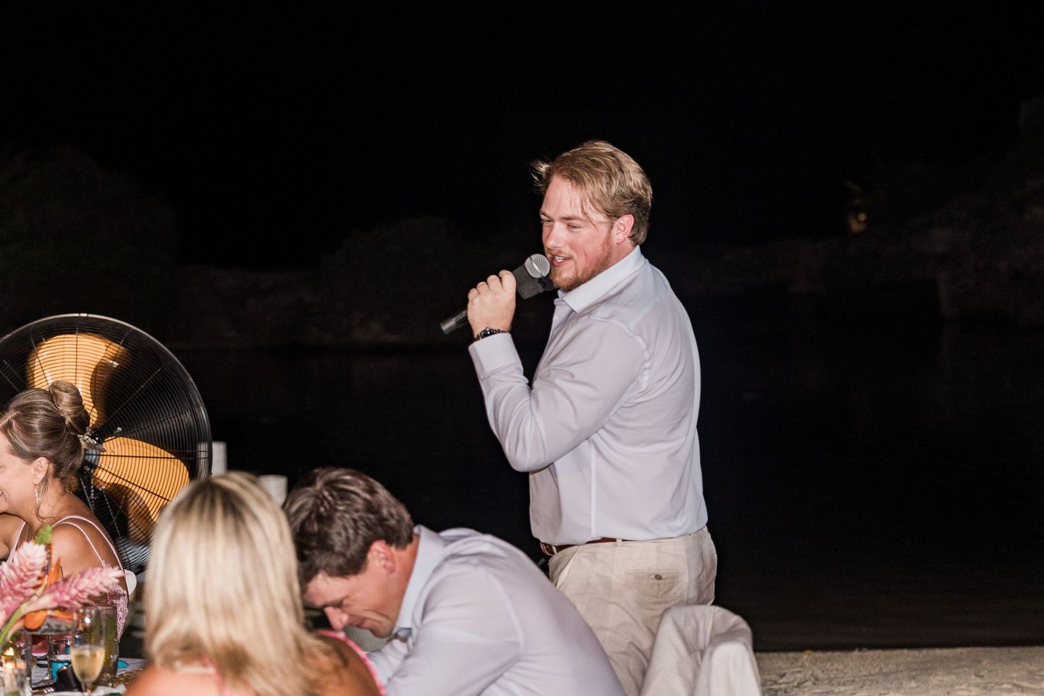 Man in gray shirt speaking into microphone at outdoor evening wedding reception dinner.