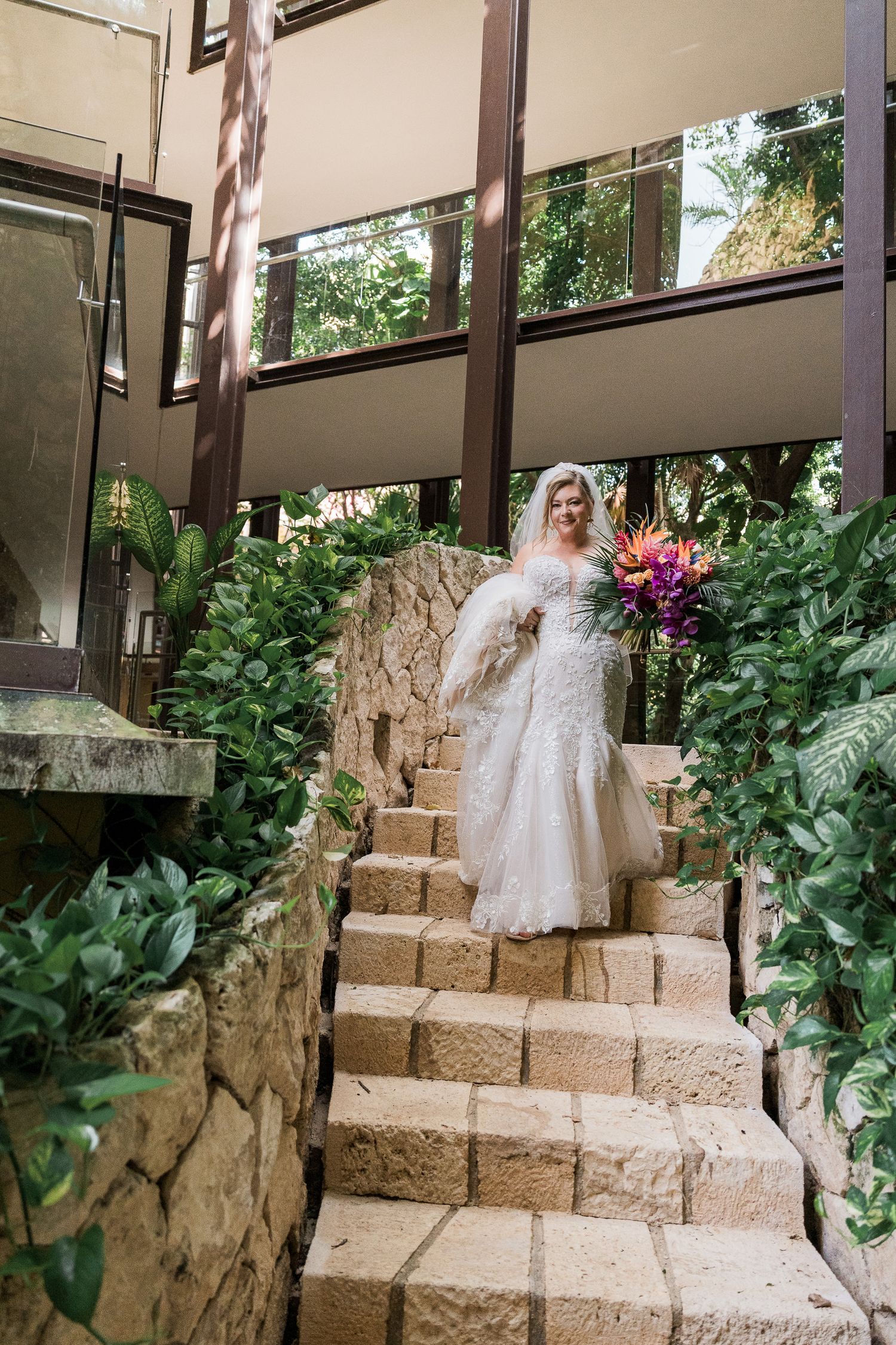 Bride in white lace gown holding tropical bouquet descends stone staircase surrounded by lush greenery at wedding venue.