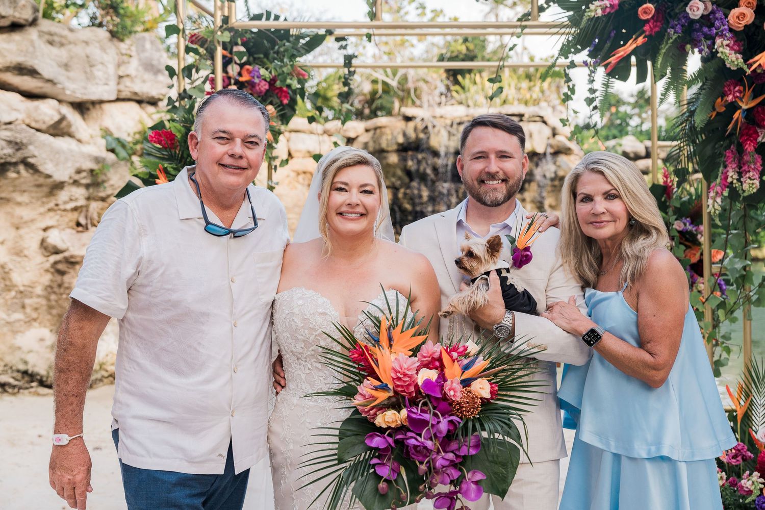 Bride and groom pose with guests holding a dog at a tropical outdoor wedding with colorful floral decorations.