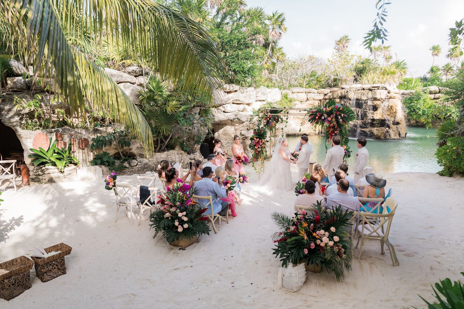 Hotel Xcaret México Intimate outdoor wedding ceremony on sand surrounded by tropical greenery, colorful flowers, and a lagoon backdrop.