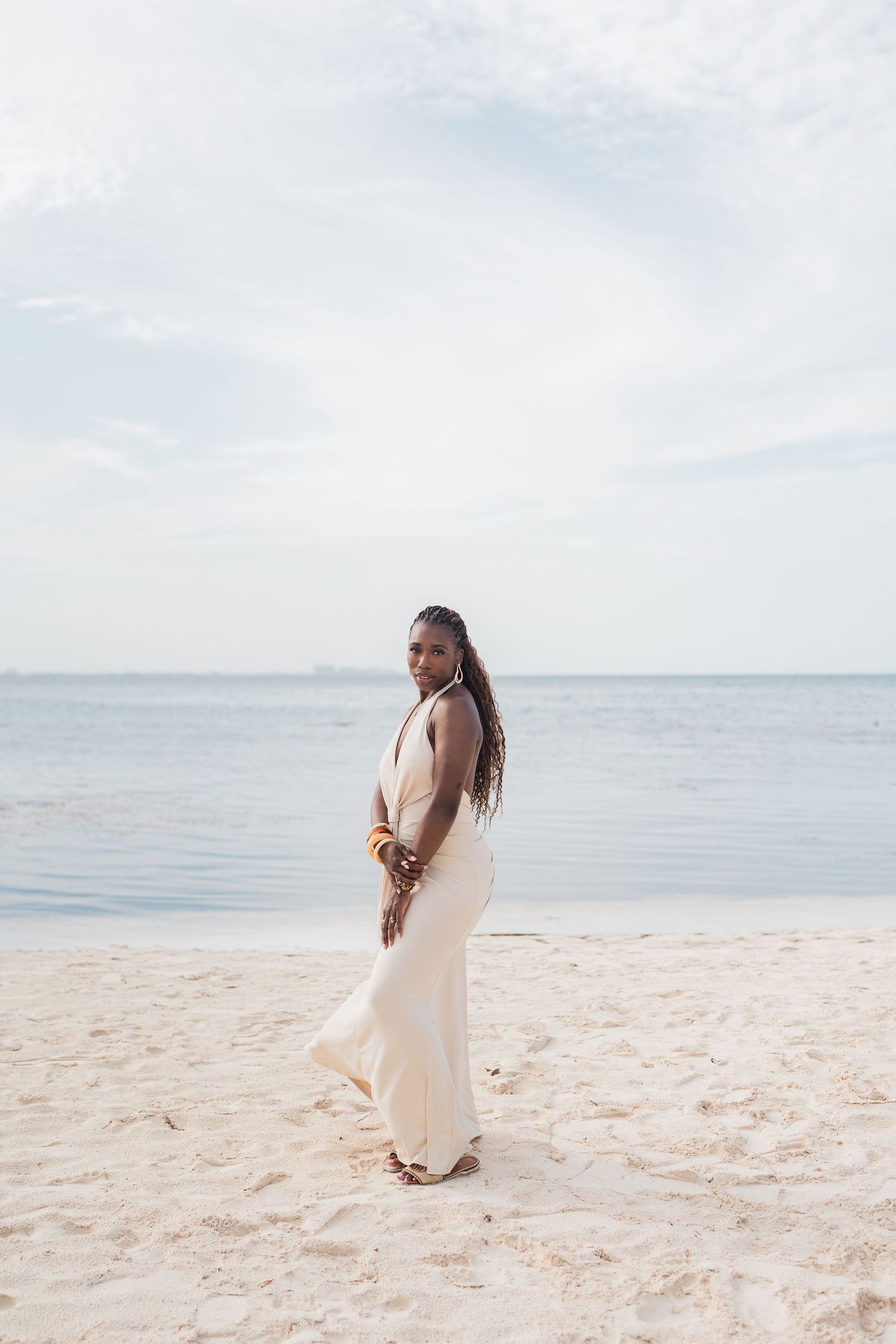 A woman in a flowing white dress stands on a sandy beach, posing gracefully with the calm ocean behind her.