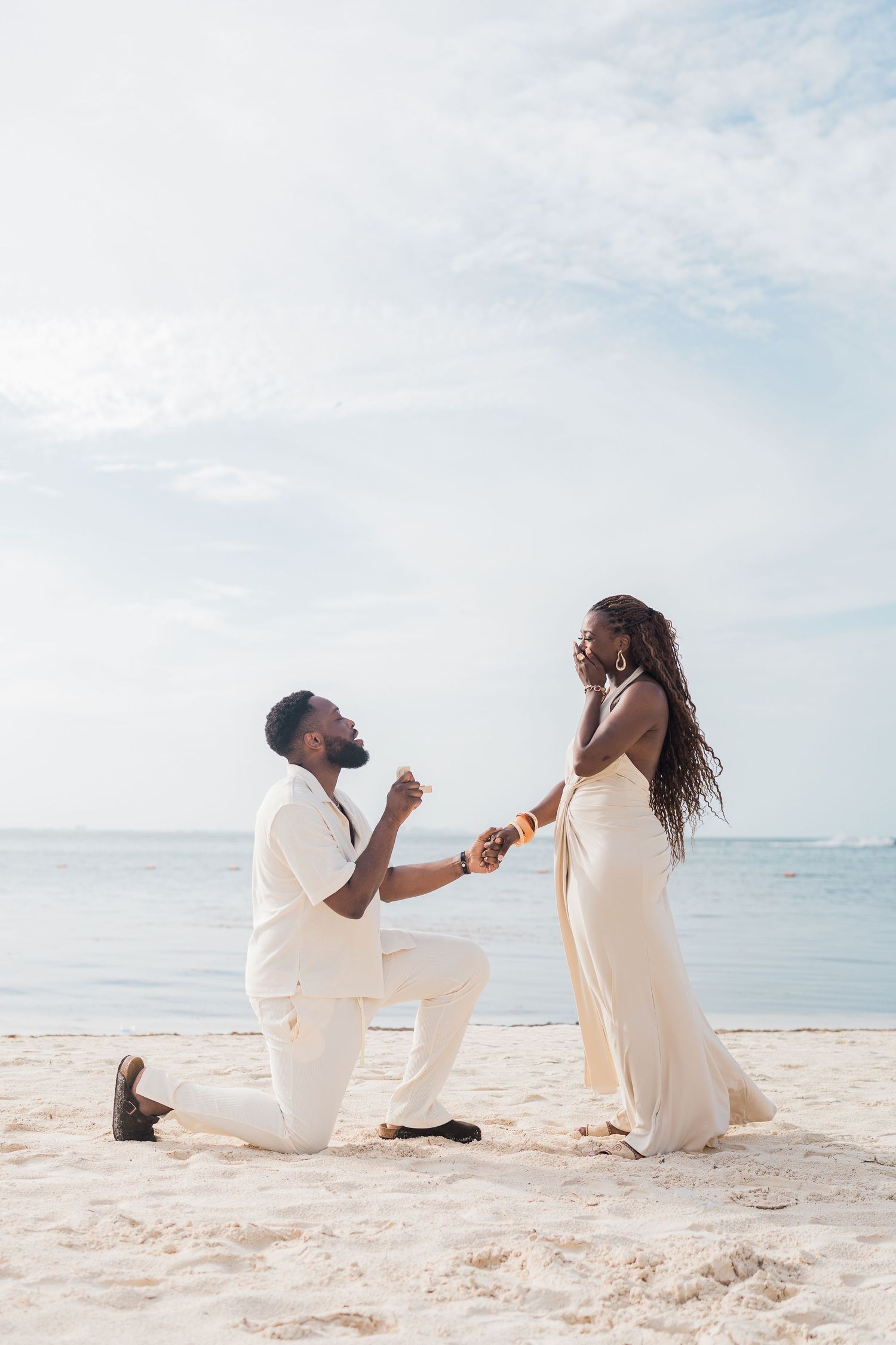 Couple in white outfits share romantic beach proposal moment by the ocean on sandy shore.