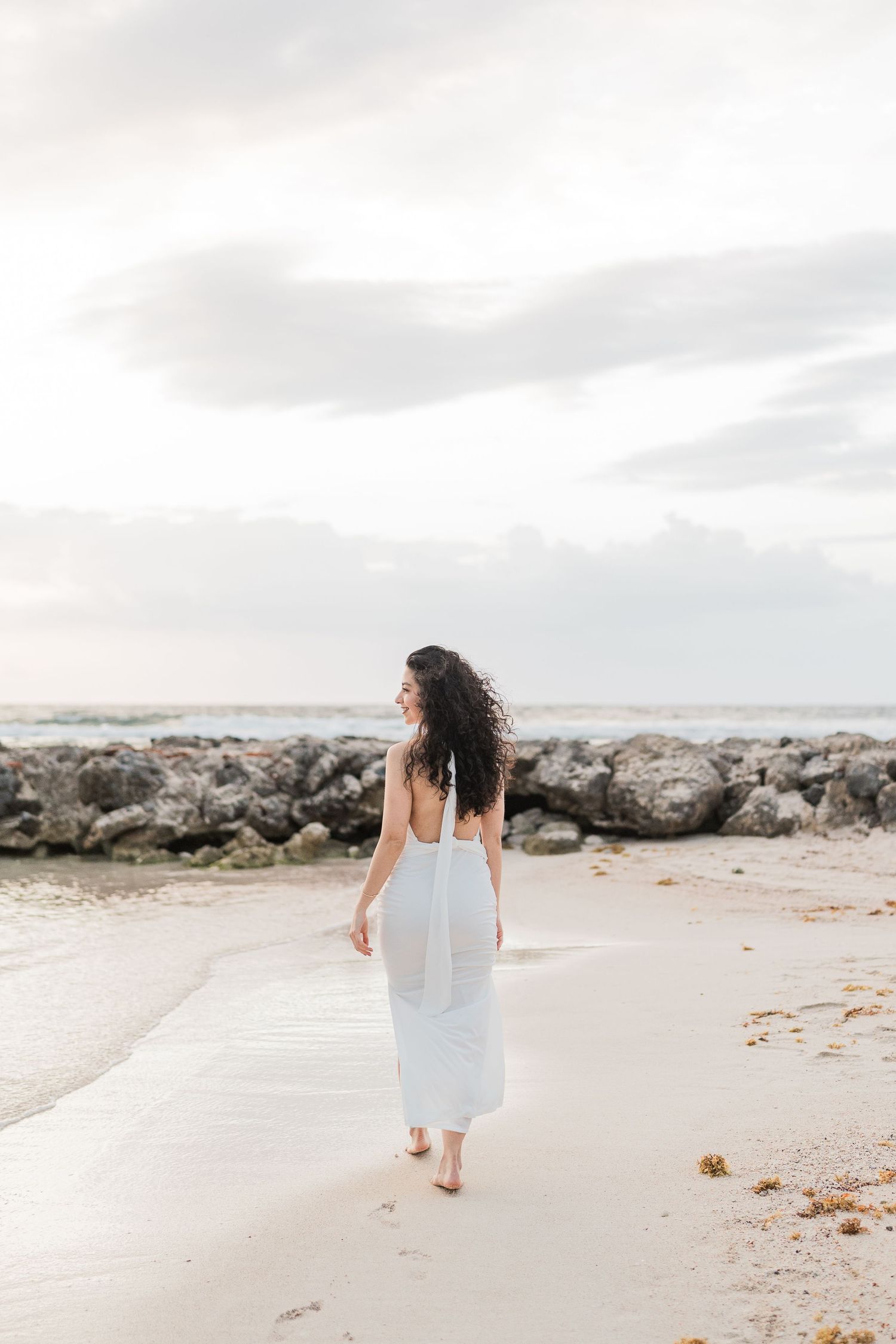 Woman in white backless dress walking barefoot on sandy beach with rocky shoreline and cloudy sky.