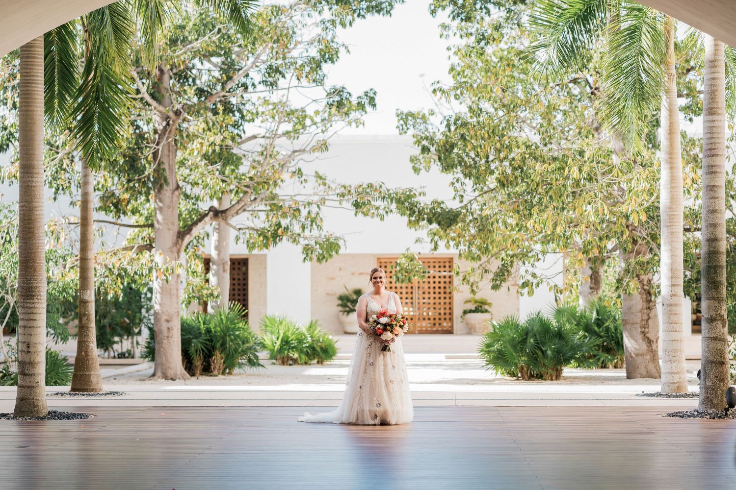 Secrets Akumal Wedding - Bride in white gown holding bouquet stands between palm trees in elegant outdoor courtyard .