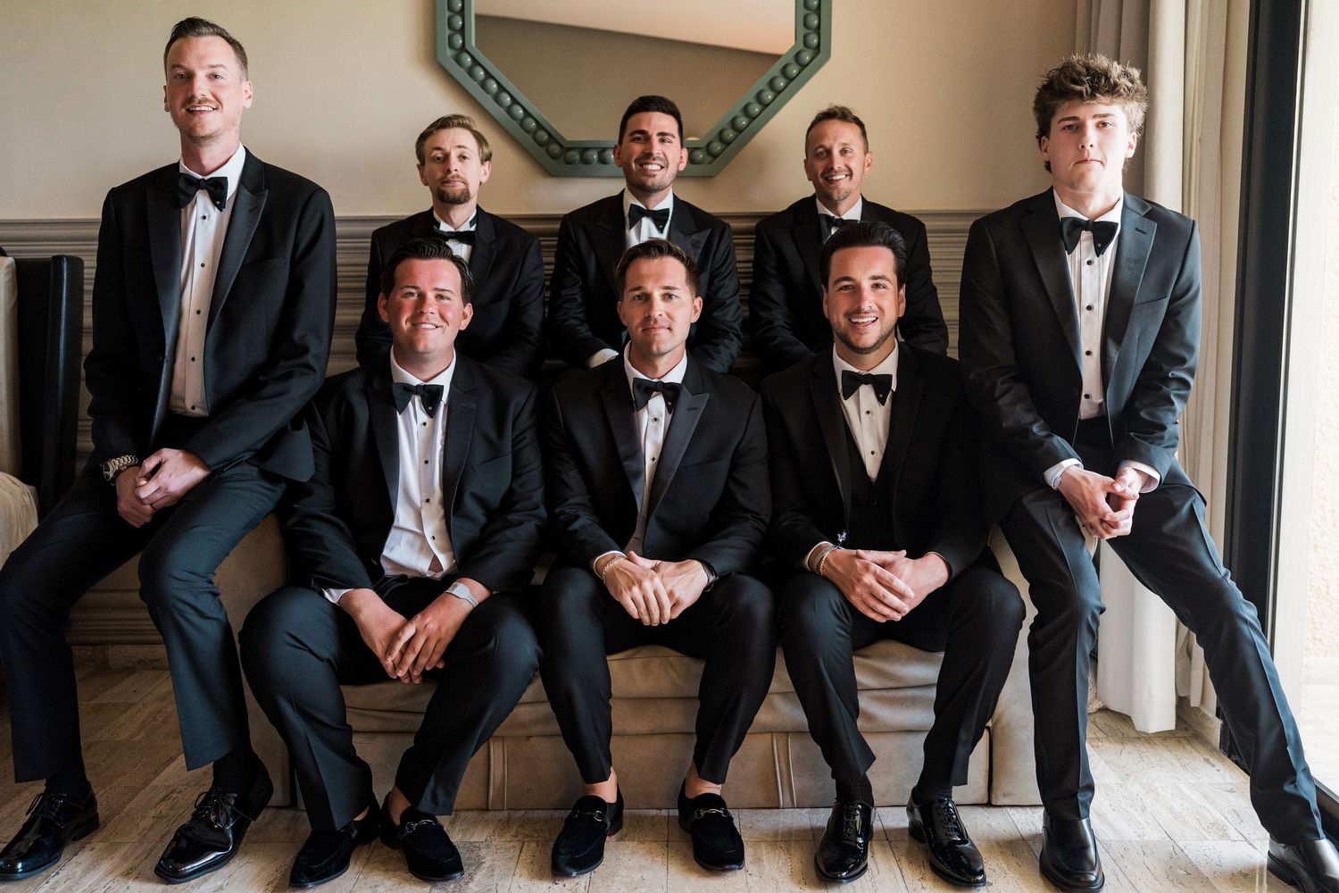Seven groomsmen in black tuxedos and bow ties pose together, three seated in front and four standing behind them.