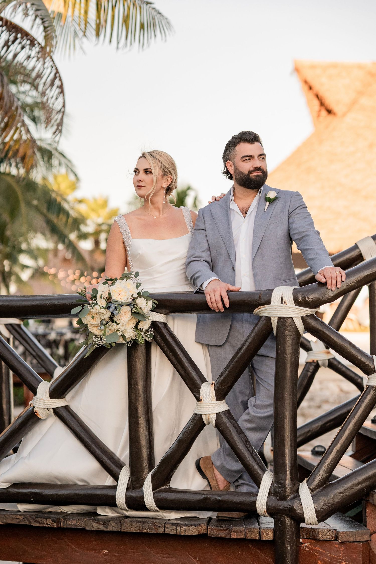 Bride and groom in elegant wedding attire stand on a rustic wooden deck surrounded by tropical palms at a resort wedding.