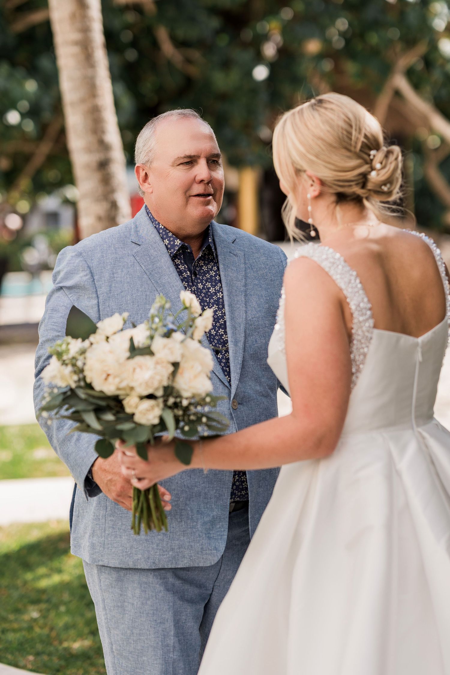 Groom in light blue suit smiling at bride in white wedding dress while holding bouquet of white roses outdoors.