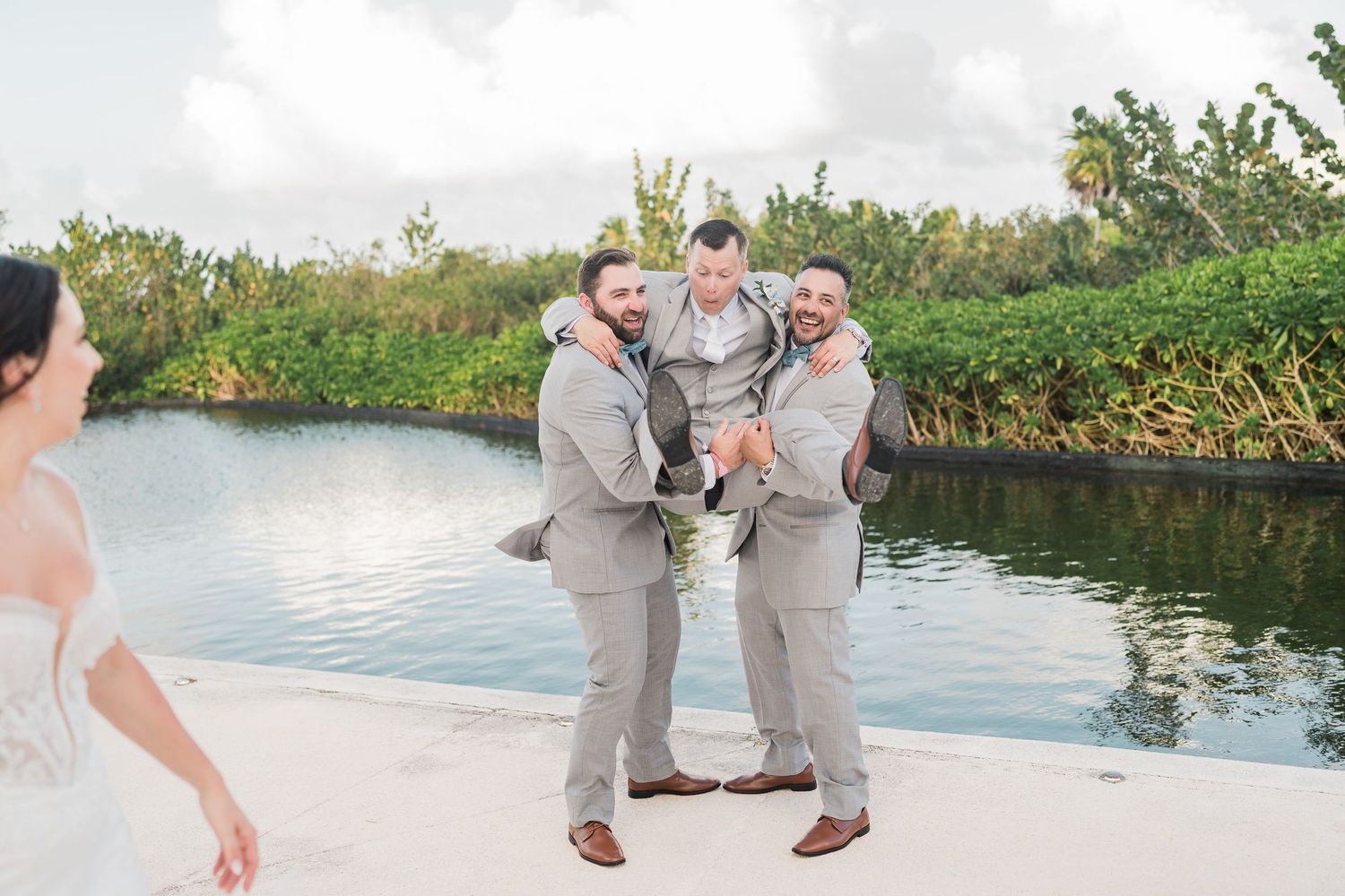 Three groomsmen in gray suits pose playfully near a waterway at an outdoor wedding venue surrounded by lush greenery.