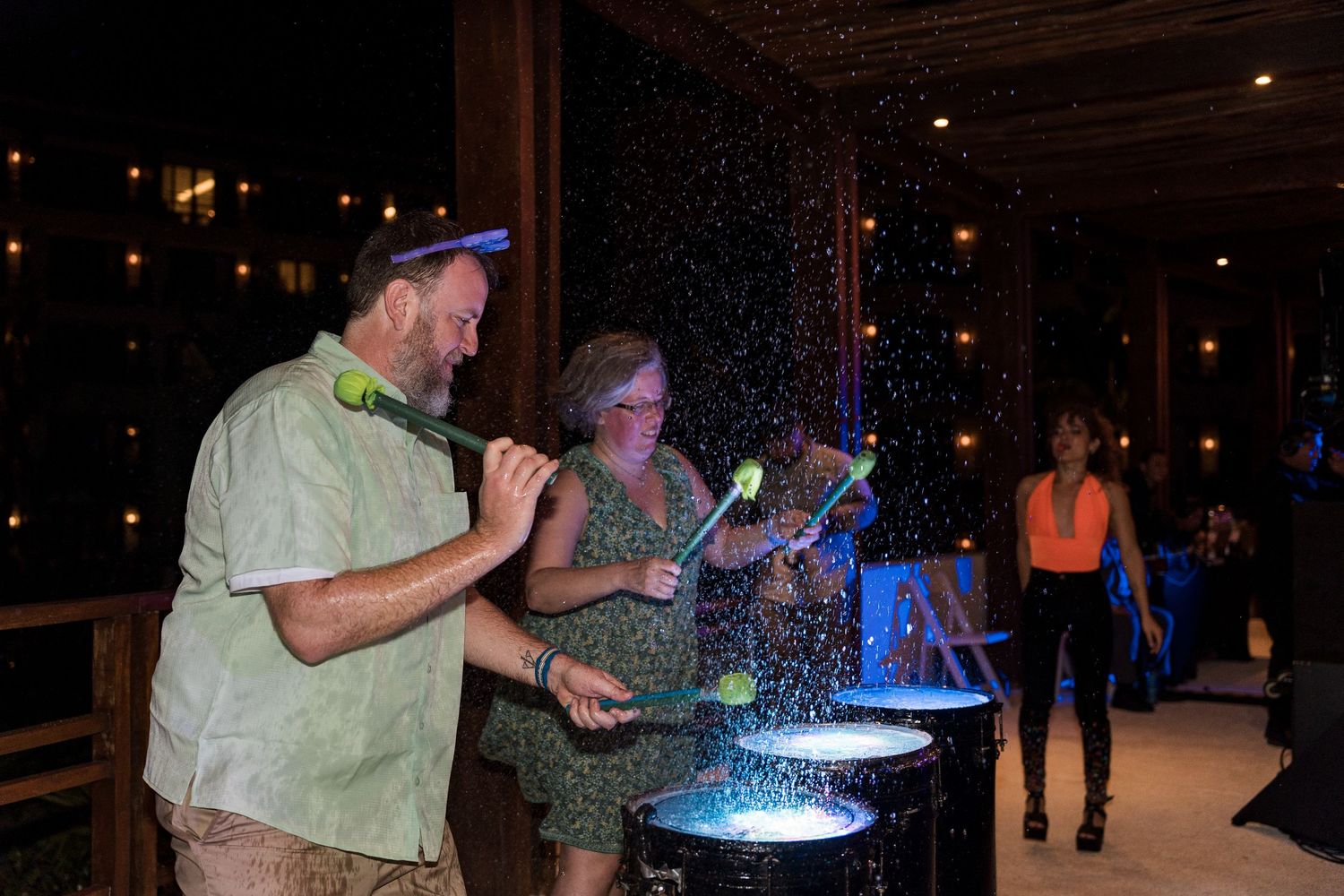 Two people playing steel drums at a lively outdoor night event with colorful lights and water splashing.