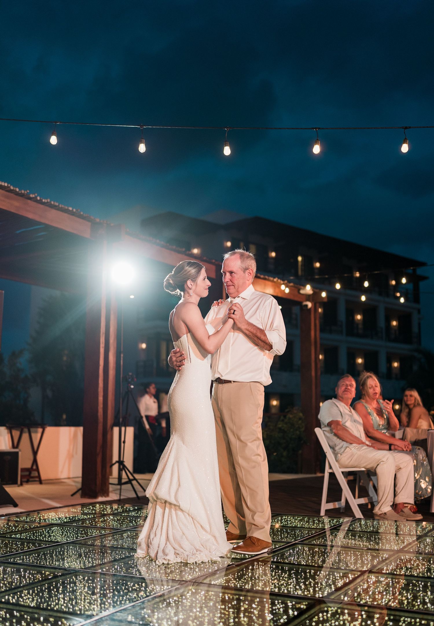 Bride and father share an emotional first dance on a mirrored outdoor floor at a romantic evening wedding reception.