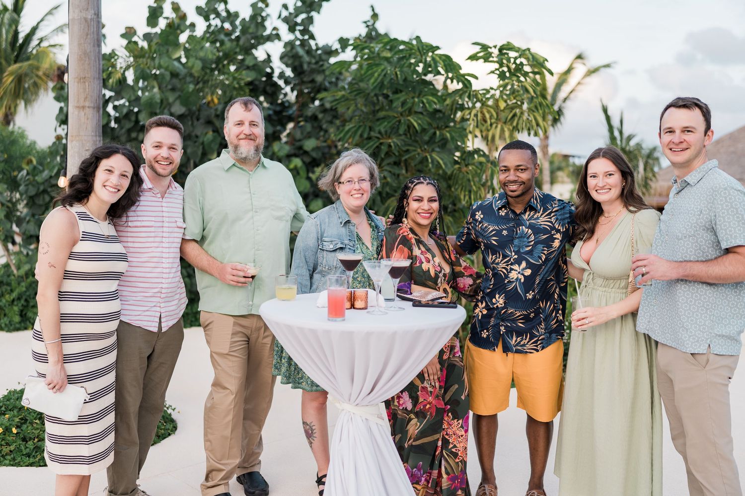 Group of eight friends smiling together at an outdoor tropical beach party with palm trees in the background.