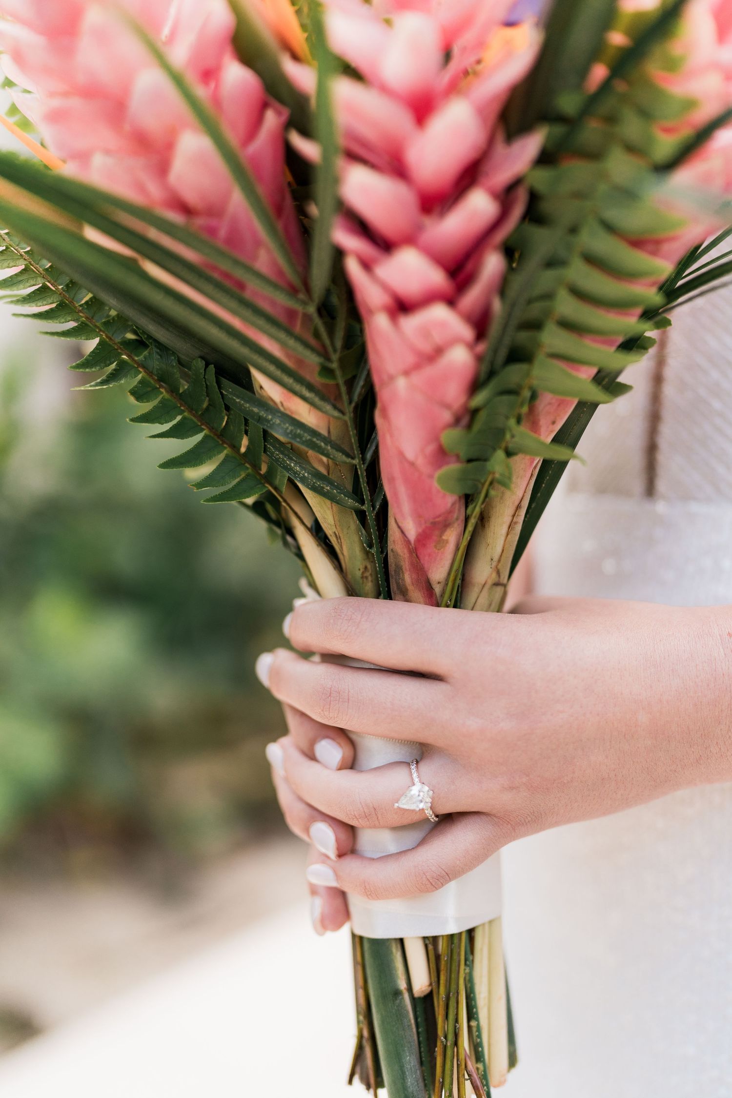 Bride holding tropical wedding bouquet with pink heliconia flowers and green fern leaves, diamond engagement ring visible.