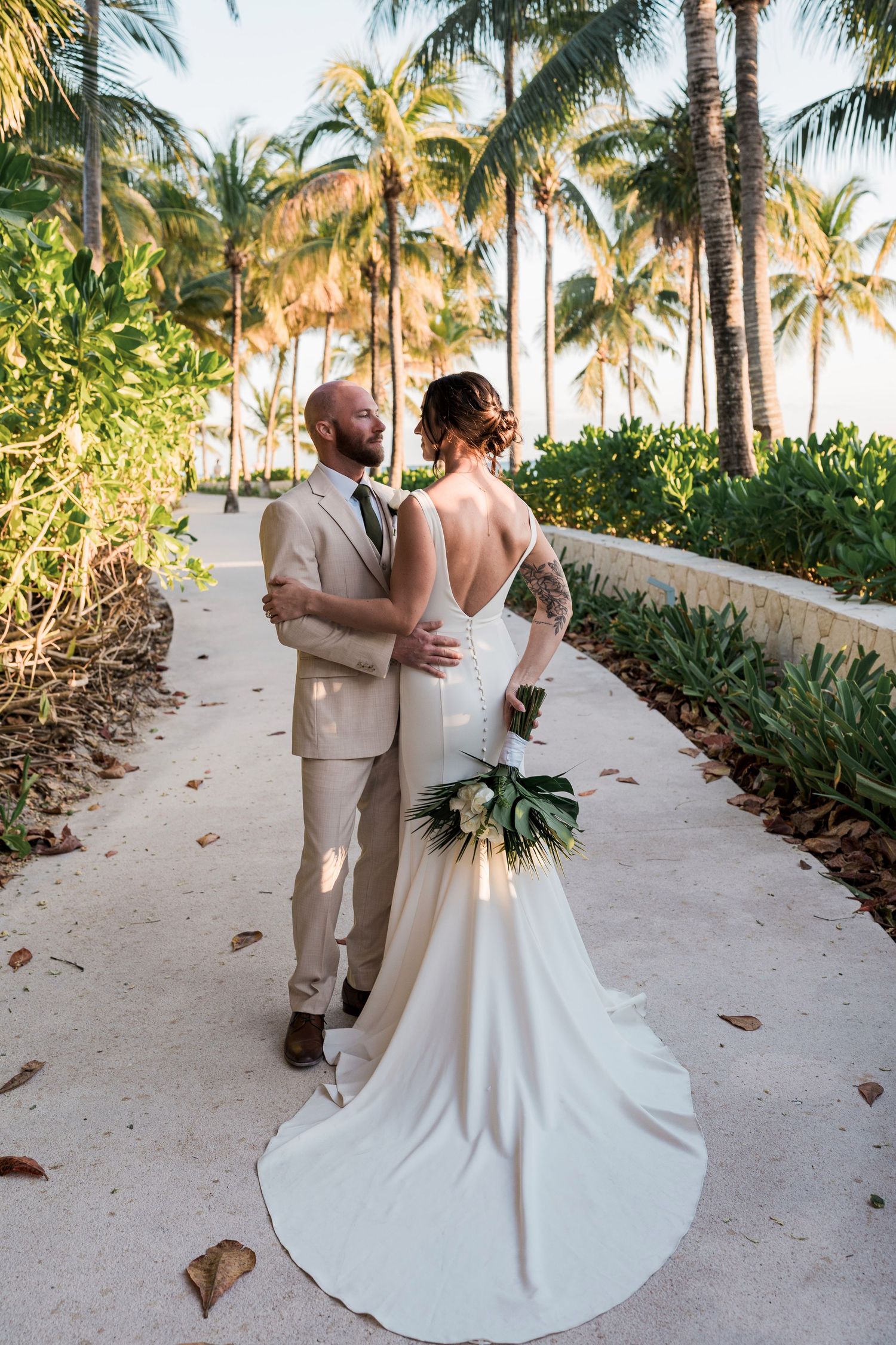 Bride and groom embrace on a tropical palm-lined path, bride in backless white gown holding bouquet at destination wedding.