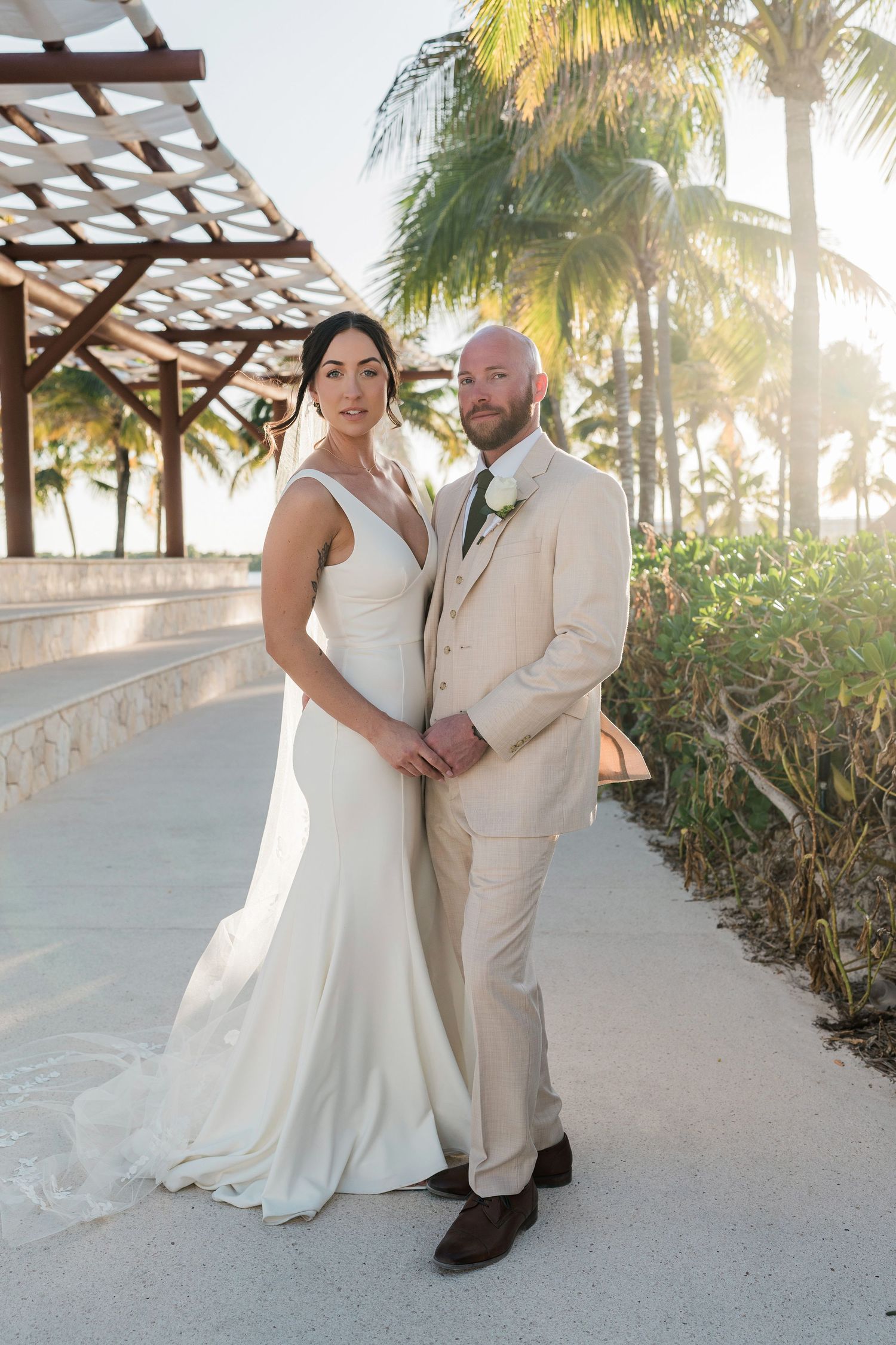 Bride in white gown and groom in tan suit pose together on a tropical resort pathway with palm trees at sunset.