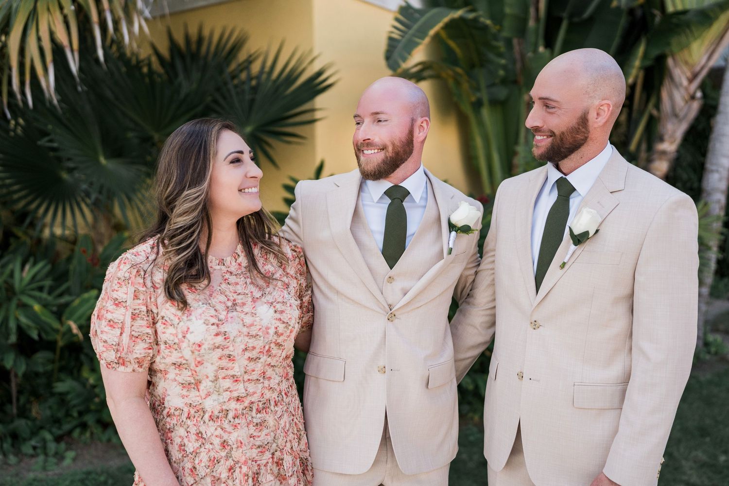 Two grooms in beige suits with green ties pose with a woman in a floral dress at an outdoor wedding celebration.