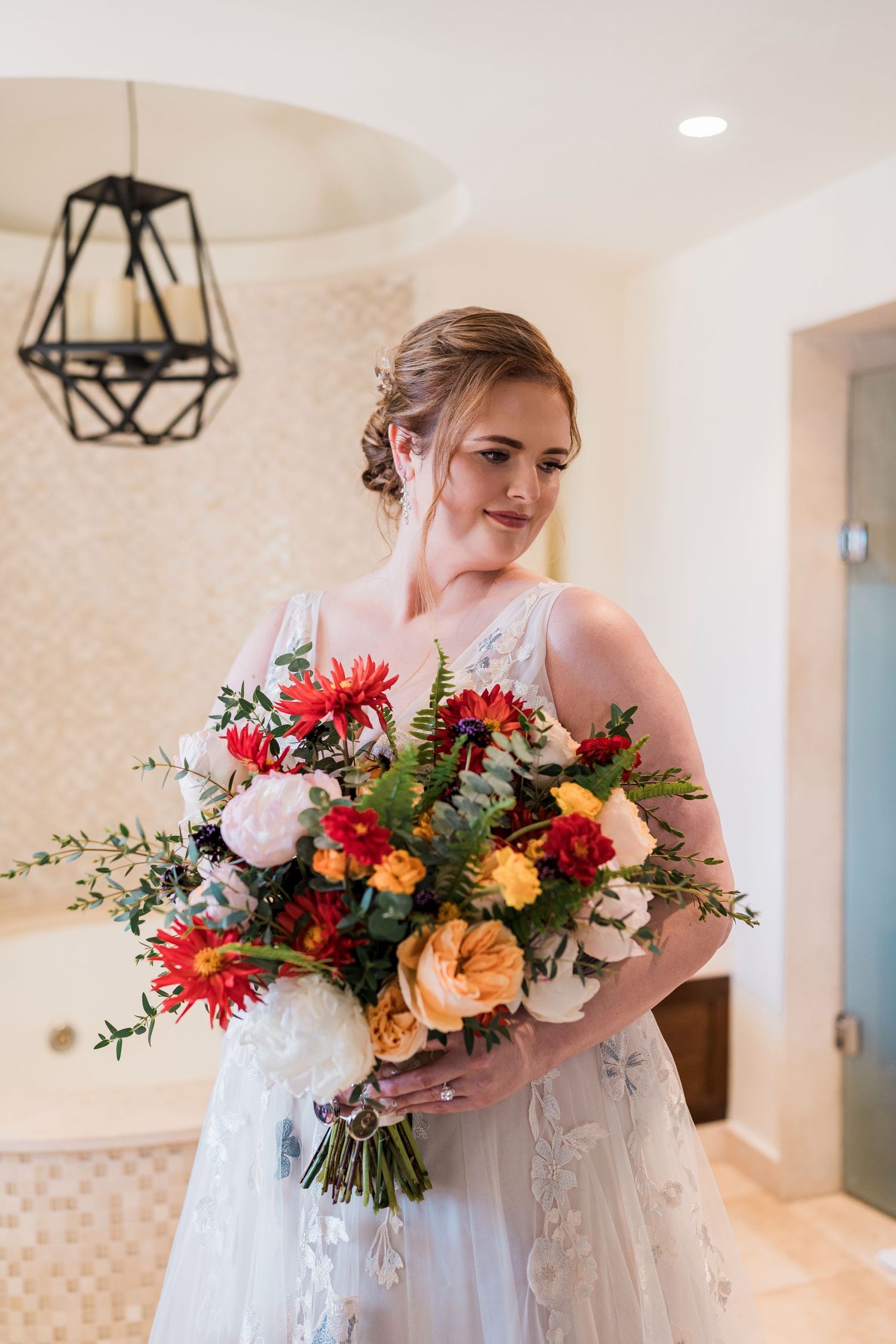 Secrets Akumal Wedding - Bride in white lace gown holding a vibrant bouquet of red, orange, and pink flowers in an elegant room.