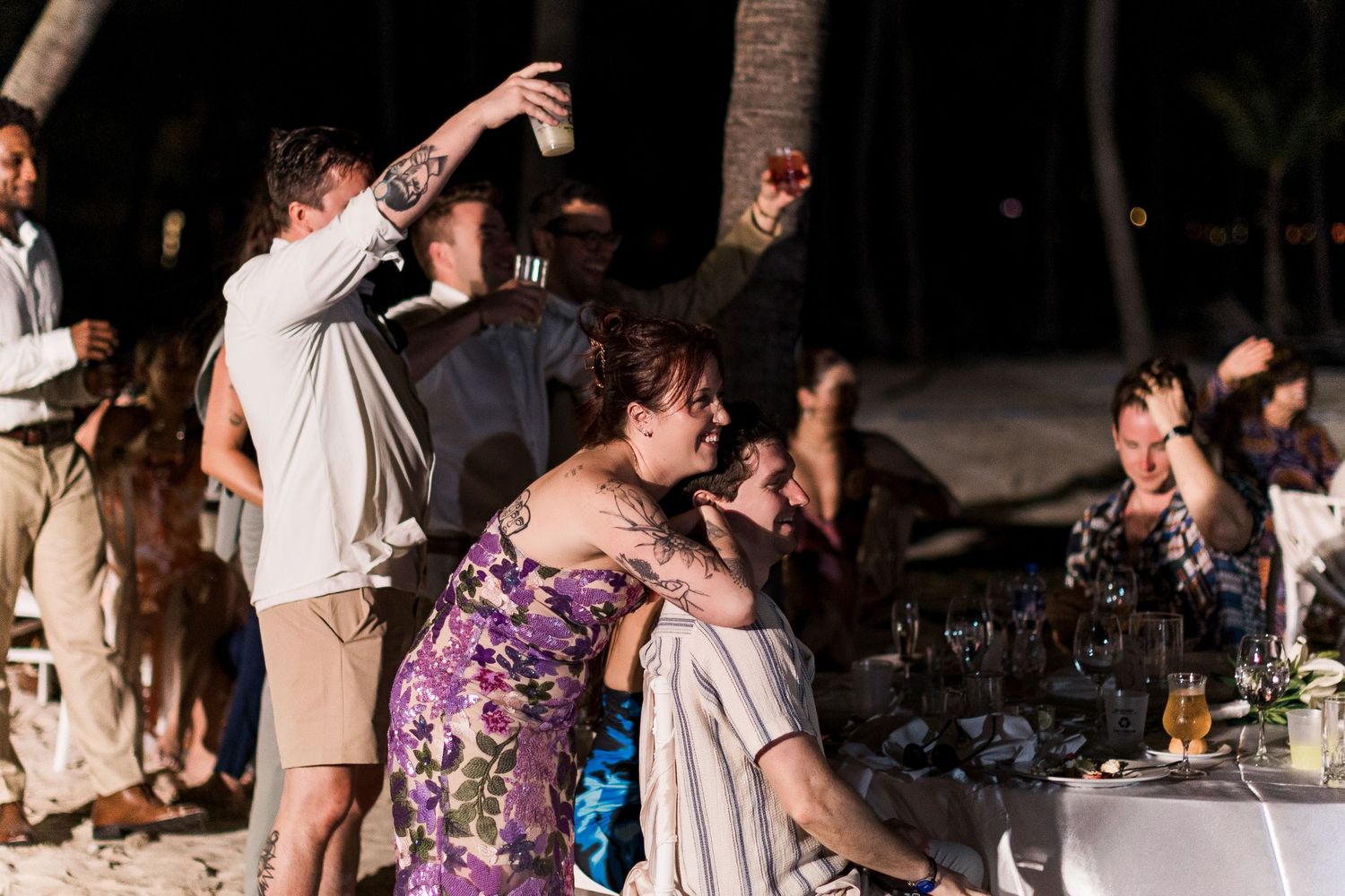 Guests celebrate at an outdoor beach wedding reception at night, raising glasses in a toast under palm trees.