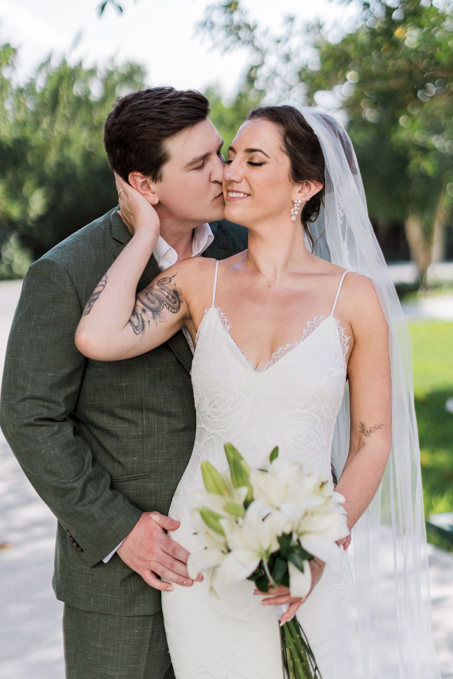 Groom in green suit kisses bride wearing white gown, veil, and holding white lily bouquet outdoors.