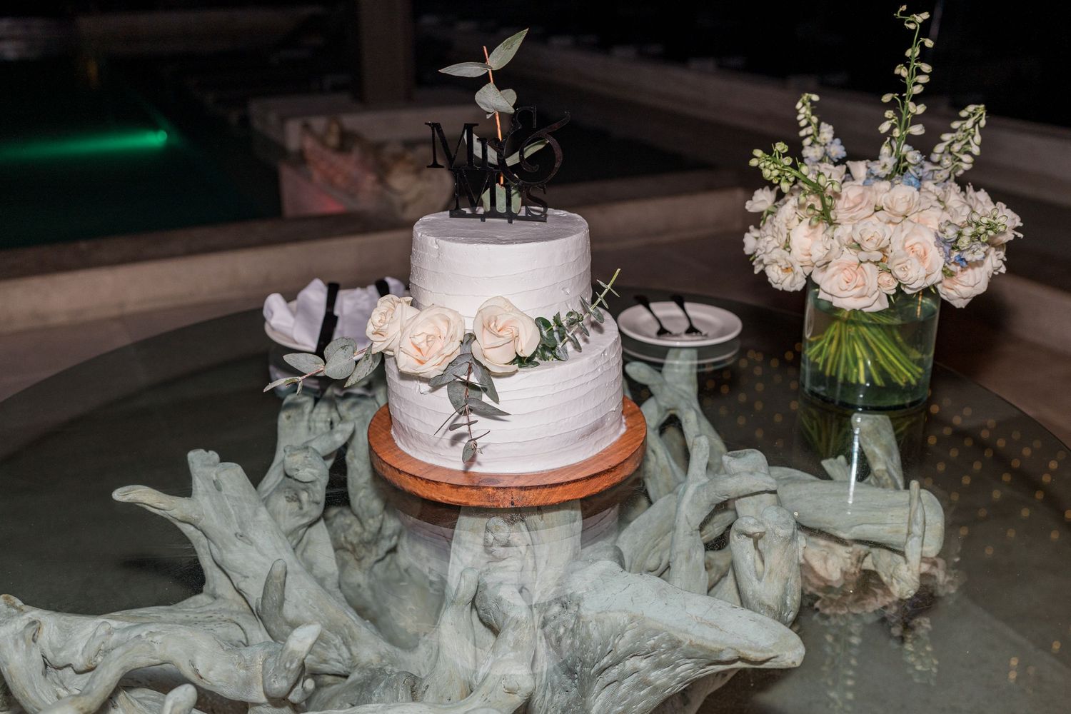 Elegant two-tier white wedding cake decorated with roses and greenery displayed on a glass table.