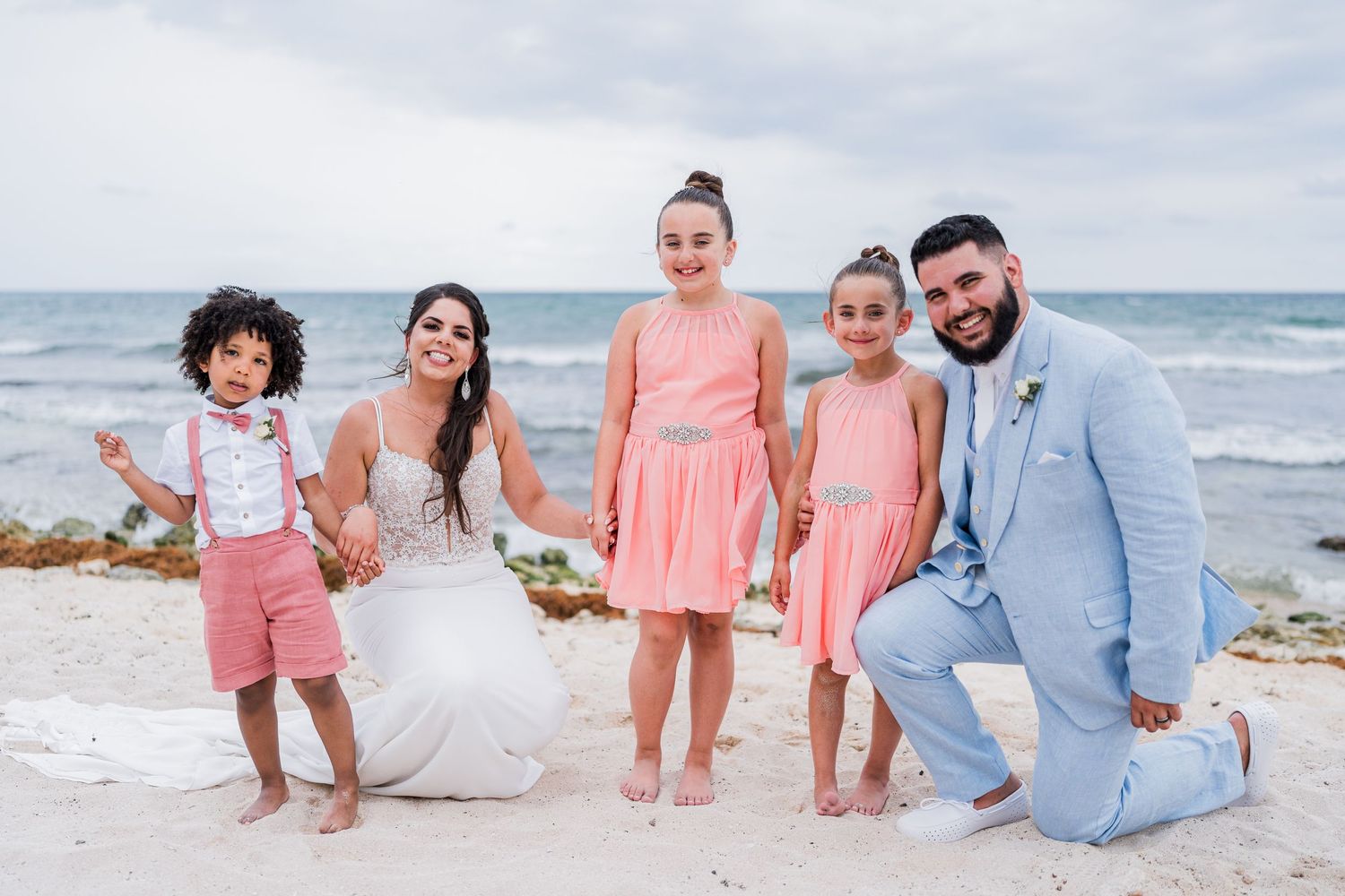 Wedding couple posing on beach with three children in coral pink outfits and a young boy in pink shorts.