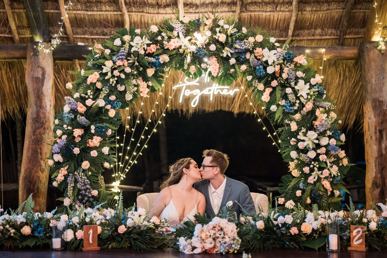 Bride and groom kiss at sweetheart table beneath floral arch with Together neon sign.