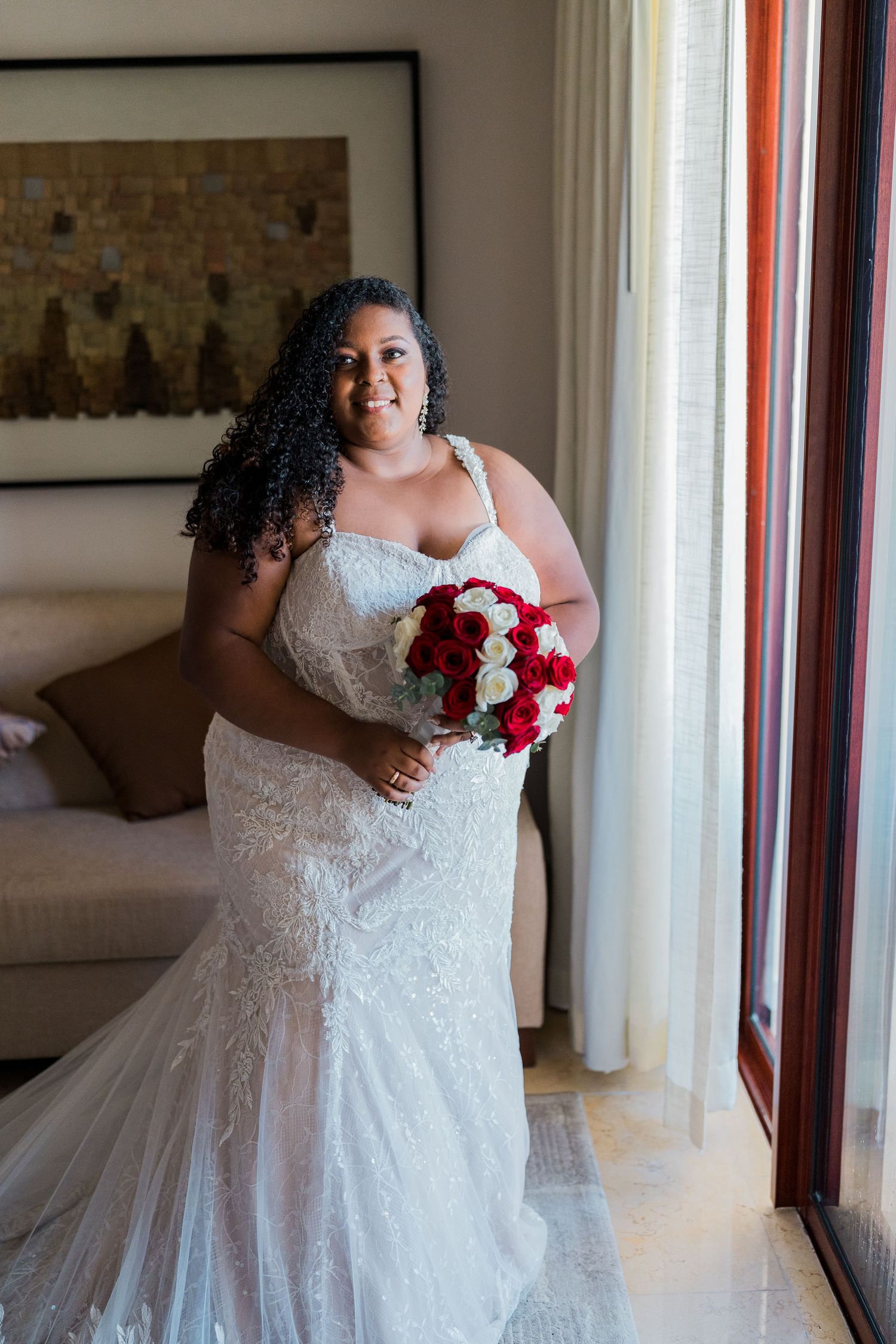Plus-size bride in white lace mermaid gown holding red and white rose bouquet by window.